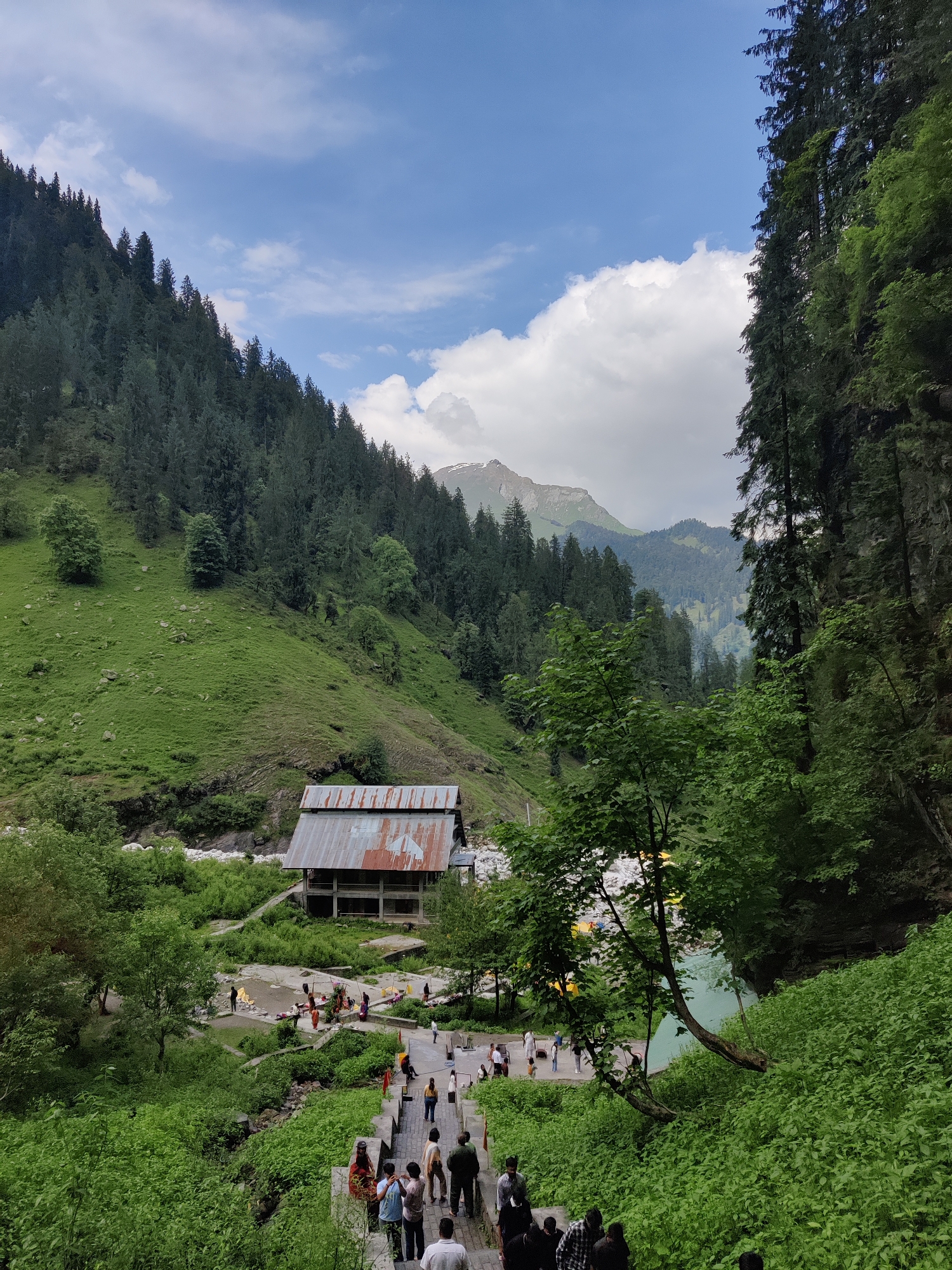 The image captures a scenic view of a mountainous landscape in the city of Majhach, India. In the foreground, a group of people are walking down a stone staircase, surrounded by lush green vegetation. Beyond the stairs, a building with a rusty metal roof stands near a flowing river. Further into the scene, rolling hills covered with dense green trees stretch towards a majestic mountain peak in the distance, under a partly cloudy blue sky. The overall impression is one of natural beauty and human presence within a serene environment.
