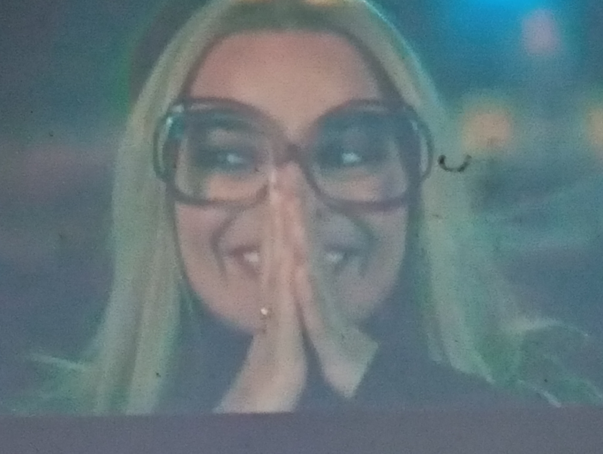 This is a close-up, slightly blurry, indoor photograph of a woman wearing large, reddish-brown framed glasses. Her hair is blonde and appears to be shoulder-length. Her hands are clasped together in front of her face, obscuring her mouth and part of her nose. She is looking towards the viewer with a happy expression, her eyes visible above the glasses. A small, sparkling earring is visible on her left earlobe. The background is out of focus and appears to be a dimly lit indoor setting with hints of blue and green light, possibly from artificial lighting or decorations. There is no visible text in the image. The image quality is low, with noticeable grain and a soft focus, suggesting it might be from an older camera or a low-resolution digital capture. The overall impression is that of a candid, joyful moment.