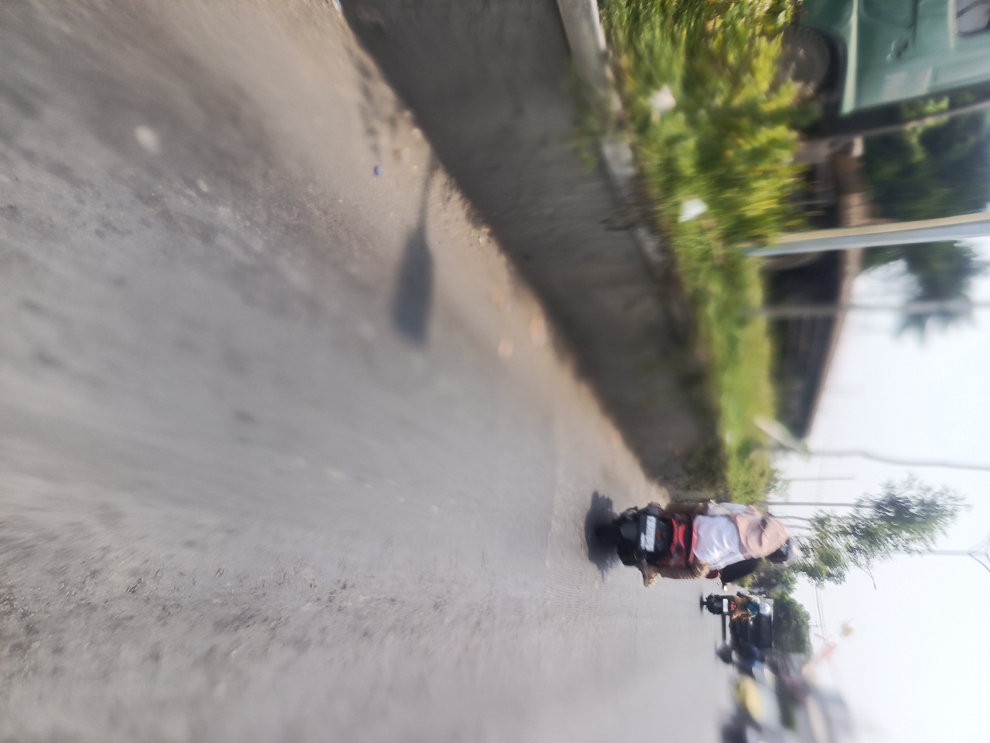 The image shows a street scene in Surabaya, Indonesia, with a focus on a motorcycle in motion. A person wearing a white shirt and a headscarf is riding the motorcycle, which has a red and black rear rack. The motorcycle is on the right side of the road, which appears to be asphalt. In the background, there are green trees and some buildings. The image is blurred due to motion, giving a sense of movement and speed. There are other motorcycles in the distance, also appearing to be in motion. The overall impression is of a busy street in an urban environment.