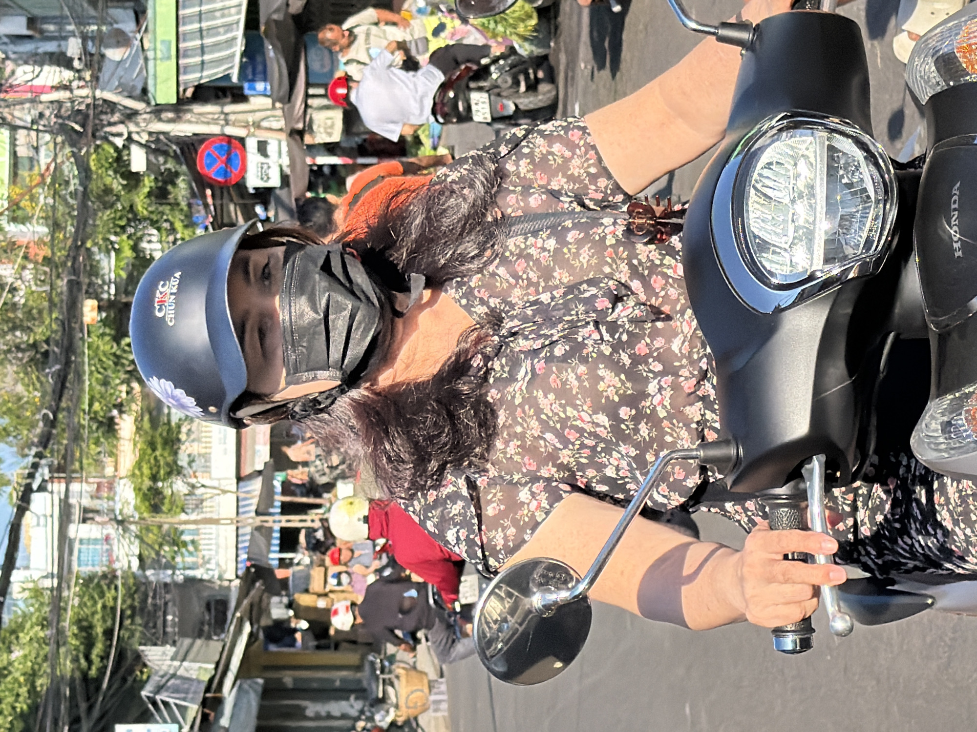 In Nha Trang, Vietnam, a woman with a black mask and a floral print dress rides a scooter. She wears a dark helmet with a flower decal and the words "CIC CHUN KUA". Her hands grip the handlebars, and the scooter's headlight is visible. Other vehicles and pedestrians are present in the background, suggesting a busy street scene. The buildings and overhead wires are characteristic of urban Vietnamese environments.