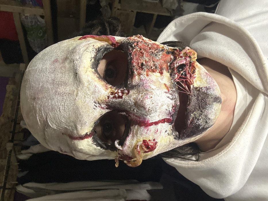 A person is depicted in a medium close-up, facing forward. Their face is covered in special effects makeup and prosthetics designed to simulate severe facial trauma and decomposition. The makeup features a pale, textured, off-white base, with darkened areas around the eyes and mouth. Dark, circular openings in the makeup reveal the individual's eyes. A prominent vertical streak of dark red, resembling dried blood, runs from the forehead down the nose bridge. The left side of the face (viewer's left) displays a yellowish, irregular protuberance near the ear and another near the outer eye corner, both with red coloration. The right side of the face exhibits extensive red, textured areas resembling exposed, torn flesh from the cheekbone to the jawline, containing small, raised, irregular elements. The mouth area is dark, mottled, and stylized to appear partially open, revealing a deep purple-black to greenish-black interior. On the right side of the mouth, thin, light-colored strands are visible across exposed red tissue. The individual wears a white hooded sweatshirt. The background is dimly lit and appears to be an indoor space, showing a clothes rack with garments on the left and wooden shelving with indistinct items on the right. This scene is located in the city of Saratov, Russia.
