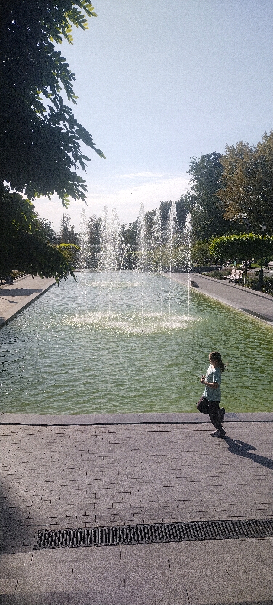 A young girl with dark hair, wearing a light blue t-shirt and dark pants, runs along a paved walkway next to a large rectangular fountain. The fountain has multiple jets of water spraying upwards. The walkway is made of grey cobblestones, and a drainage grate runs along the edge closest to the camera. In the background, trees line the perimeter of the fountain, and a clear blue sky is visible. The scene appears to be in a park or public square. This image was captured in Kharkiv, Ukraine.