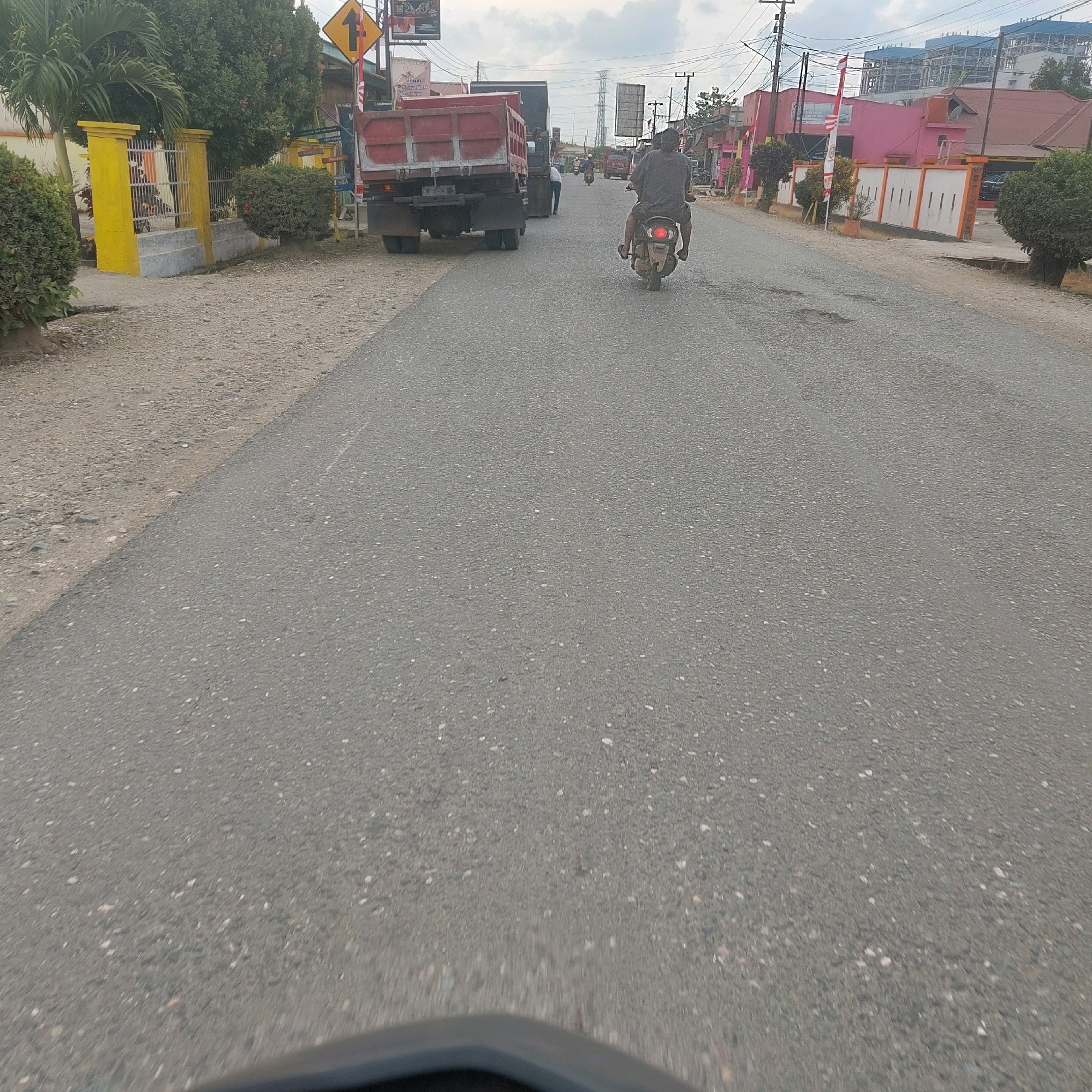 The image depicts a street scene in Topogaro, Indonesia. A person is riding a motorcycle on an asphalt road, with a red dump truck parked on the side of the road. The truck has a license plate that is partially visible. Several buildings line the street, including a pink building with white fencing and a building under construction. There are also trees, power lines, and traffic signs present. The road appears to be in moderate condition with some potholes. The sky is overcast.