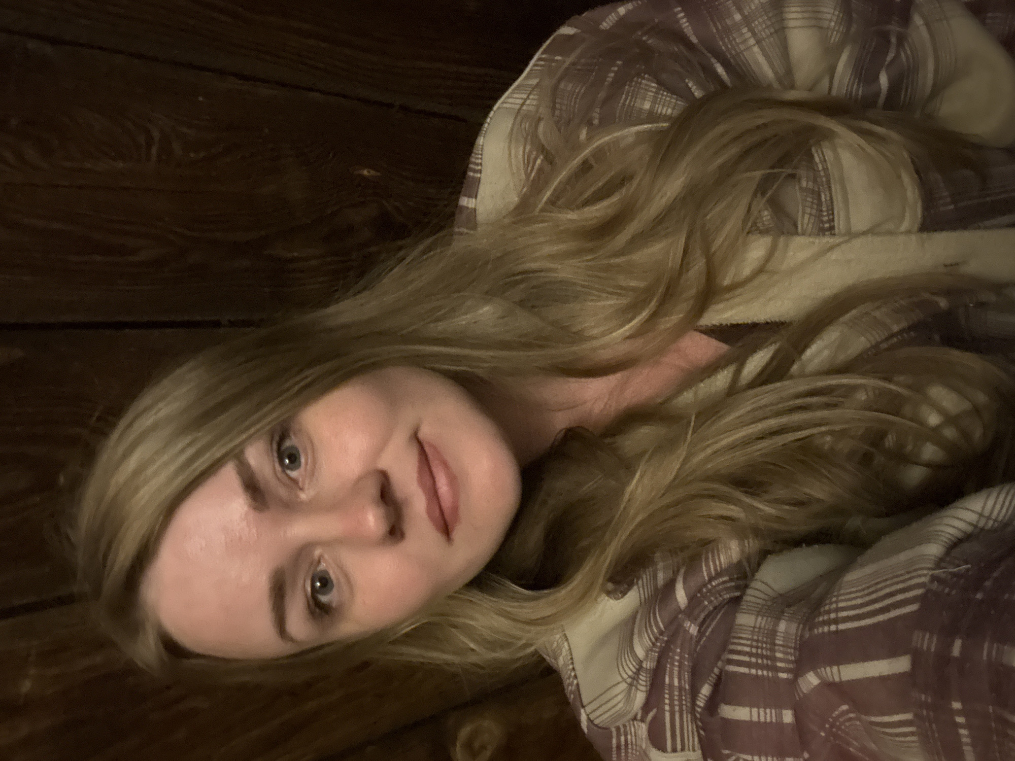 A blonde woman is laying down on what appears to be a wooden surface. She is wearing a brown and white plaid shirt. It is impossible to determine her exact location in Chaikovsky, Russia from the given image.