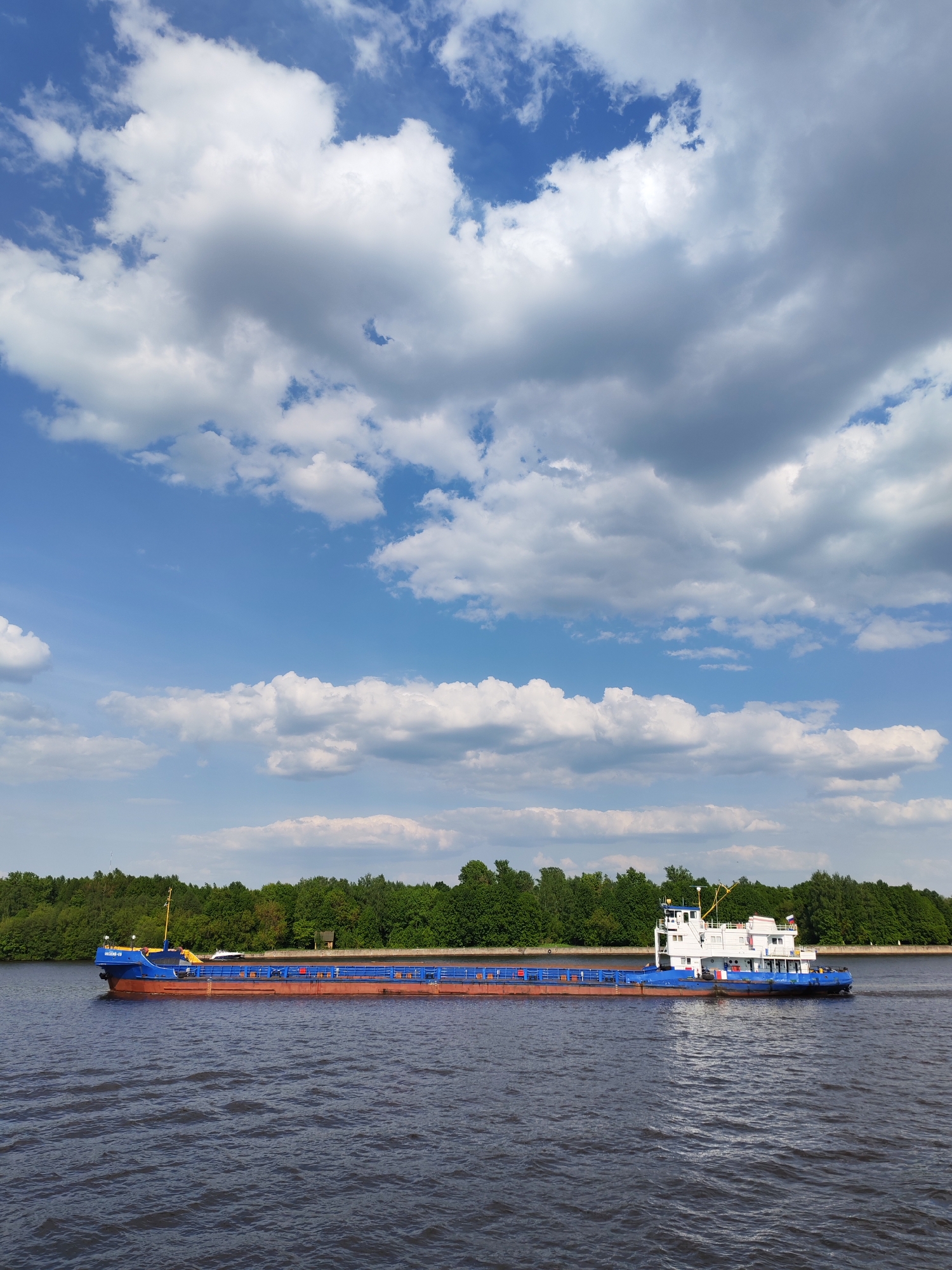 A blue and red barge navigates a wide river in Ermolino, Russia. The boat's hull is blue at the front and back, with an orange-red center section. A white superstructure sits atop the rear of the barge. The water is dark and choppy, with subtle ripples indicating movement. A strip of green trees lines the far shore. Above, a bright blue sky is filled with puffy white clouds. The scene is bright and sunny, indicating a clear day.
