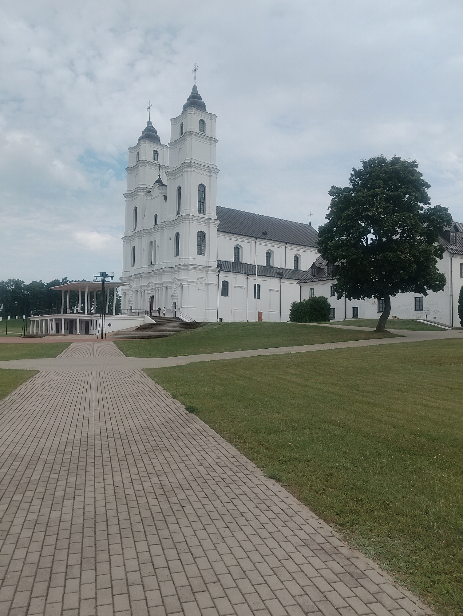 The image depicts a view of the Basilica of the Assumption of the Blessed Virgin Mary in Aglona, Latvia. In the foreground, a paved walkway made of grey interlocking pavers extends into the distance, bordered by a well-maintained green lawn. To the right of the walkway, a large, mature tree with dark green foliage stands prominently.
The basilica itself is a prominent white building with a dark roof. It features two symmetrical bell towers, each topped with a black spire and a cross. The architecture is classical and imposing, with arched windows and decorative elements along the facade. To the left of the basilica, an open-air pavilion with a white roof and supporting pillars is visible, with steps leading up to it. Two individuals are seen walking up the steps towards the basilica entrance, and a few other figures are present on the grounds near the pavilion.
The sky is overcast with a mix of grey and white clouds, suggesting a temperate day. The surrounding area appears to be a park or landscaped grounds, with trees visible in the background to the left. The overall scene conveys a sense of tranquility and architectural grandeur.