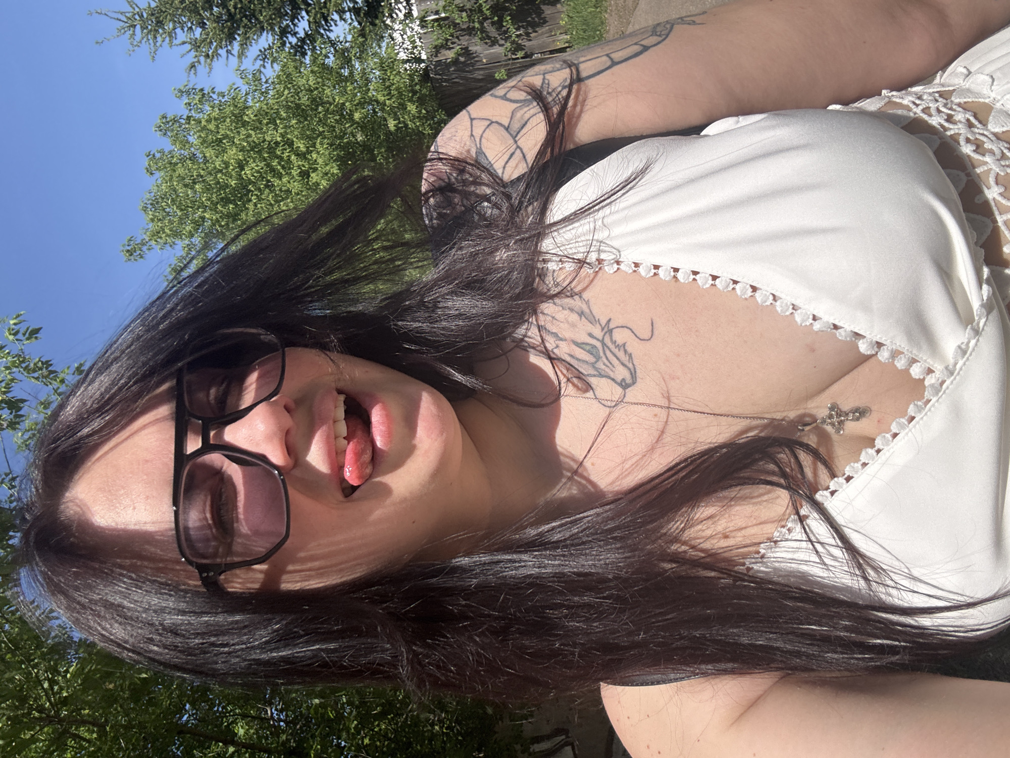 A woman is seen in an outdoor setting. She has long, dark hair and is wearing sunglasses. Her tongue is sticking out. She is wearing a white top with lace detailing and a silver necklace with a cross pendant. Tattoos are visible on her arm and chest. The background includes trees and a blue sky. The location is not determinable based on the provided information.
