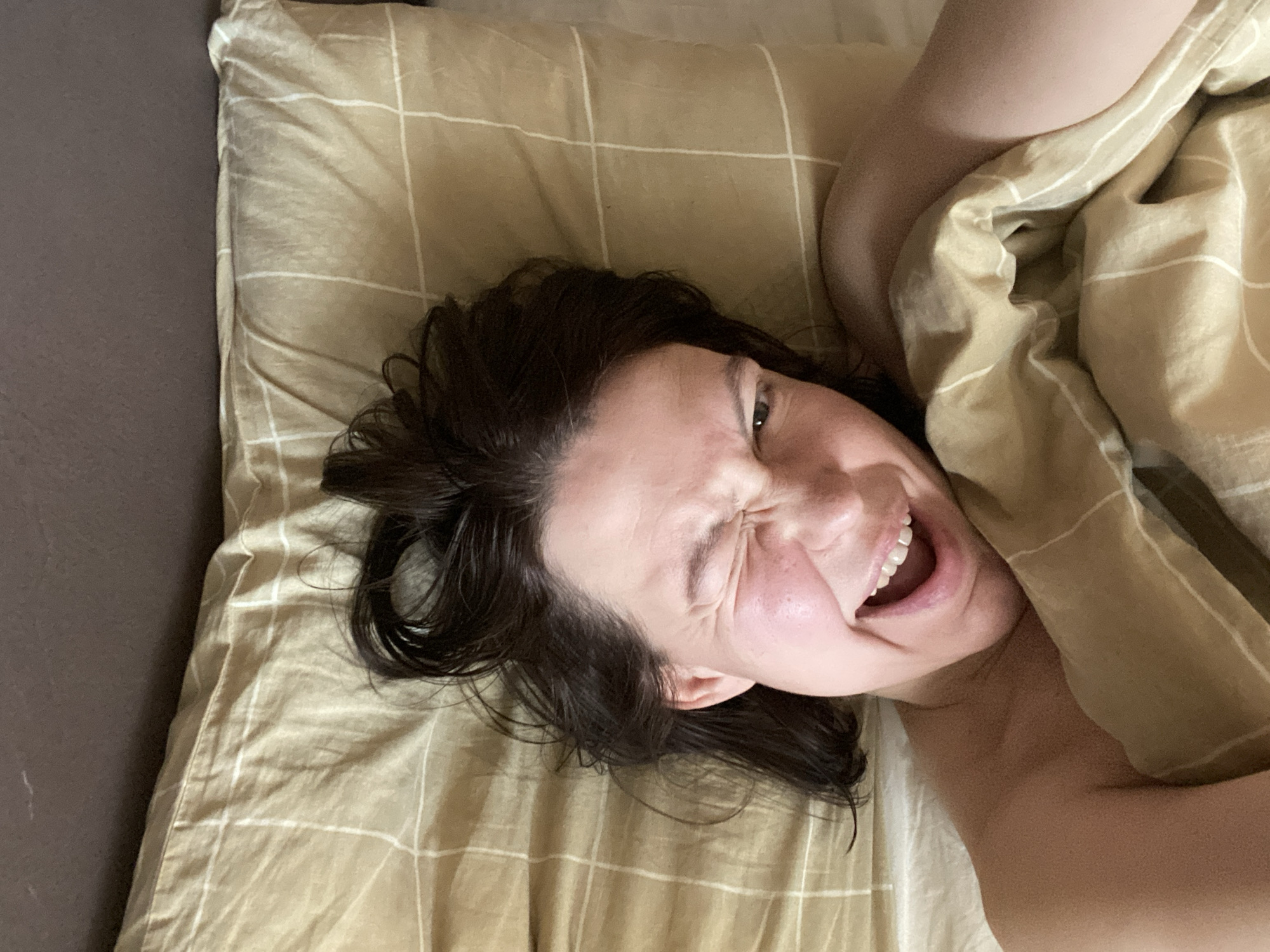 An overhead shot captures a person lying on a bed in an indoor setting. The individual, appearing to be an adult female with dark, slightly disheveled hair, occupies the central and right portions of the frame. Her head rests on a light brown or beige pillow, which features a subtle white grid pattern. To the left of the pillow, a plain dark brown surface, likely part of the bed or mattress, is visible.
The person is partially covered by a matching light brown or beige blanket or duvet, also adorned with a white grid pattern, which drapes over her right shoulder and arm. Her face is turned slightly towards the camera, and she displays a highly animated and expressive emotion. Her eyes are tightly scrunched, and her mouth is wide open, revealing her teeth, in what appears to be a laugh, a shout of joy, or perhaps a playful scream. This suggests a moment of intense happiness, amusement, or excitement. The setting is a generic domestic bedroom, and there are no specific visual cues within the image to indicate the time of day, weather, or the provided location context of Chaikovsky, Russia. No visible text is present in the image.