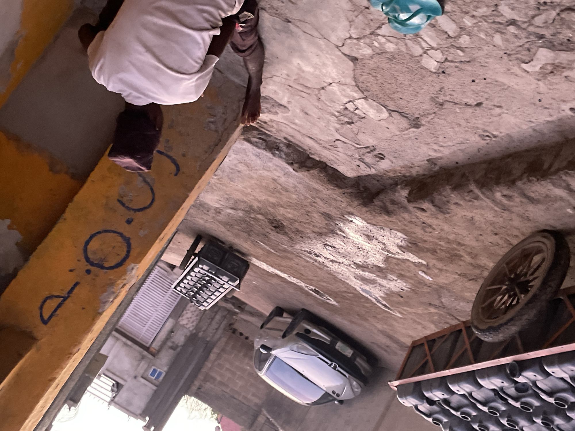 The image captures an outdoor scene, likely a narrow alleyway or utilitarian space, from a high, tilted perspective. The ground is a patchwork of cracked and uneven concrete.
In the foreground, a person is bent over, facing away from the camera. They wear a white short-sleeved shirt and dark trousers, with bare feet visible on the worn concrete. Their head is obscured, suggesting they are focused on something on the ground or engaged in an activity requiring a low posture.
To the immediate left, a bright yellow architectural element, possibly a beam or a low wall, runs diagonally through the frame. Painted on this yellow surface are blue characters that read "D. O." followed by a circular shape, possibly a "0" or "O".
In the mid-ground, a black, open-grid crate or cage rests on the ground. Nearby, a silver or light-colored car is parked, its front facing away from the viewer. Further into the background on the left, parts of buildings are visible, including a small blue-framed window and a white louvered structure.
On the right side of the frame, a wooden spoked wheel is propped up or part of a structure. Below it, a wooden rack holds numerous dark-colored plastic containers or jerrycans arranged in rows. In the upper right corner, a pair of light blue flip-flops lies on the cracked pavement.
The lighting indicates daytime, but without strong shadows, it could be overcast, early morning, or late afternoon. The scene conveys an atmosphere of a lived-in, possibly working, environment.