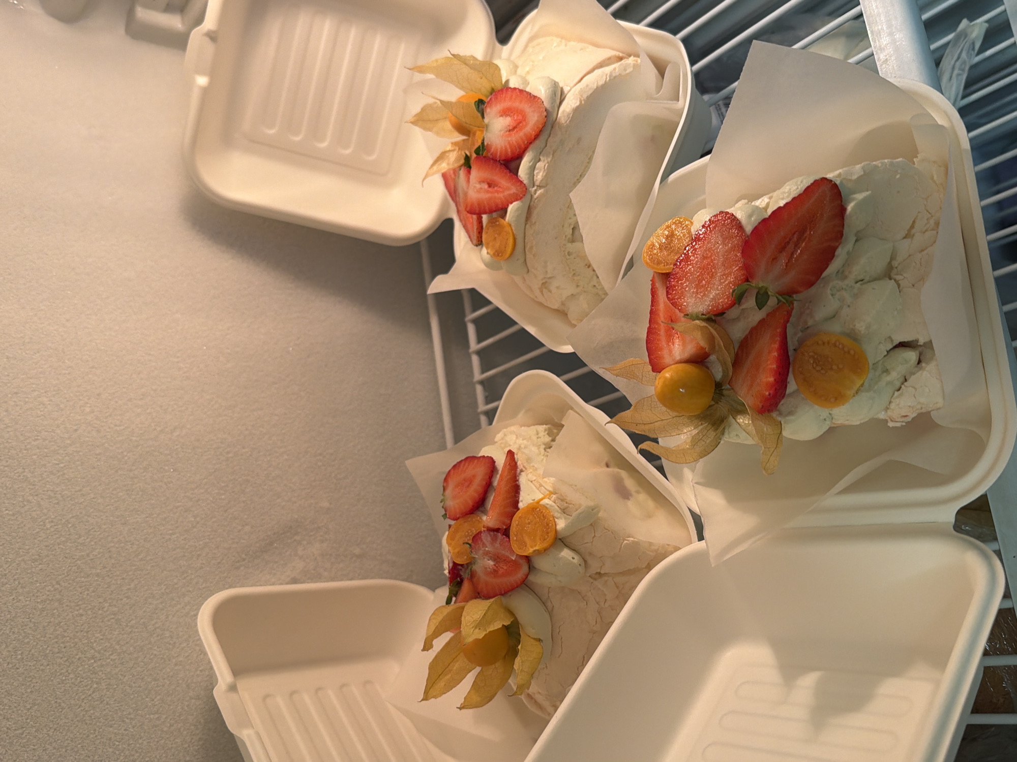 The image shows three meringue desserts topped with strawberries and golden berries in white takeaway containers. They are stacked on a wire rack, possibly inside a refrigerator. The scene appears to be indoors. The location is not evident in the image.
