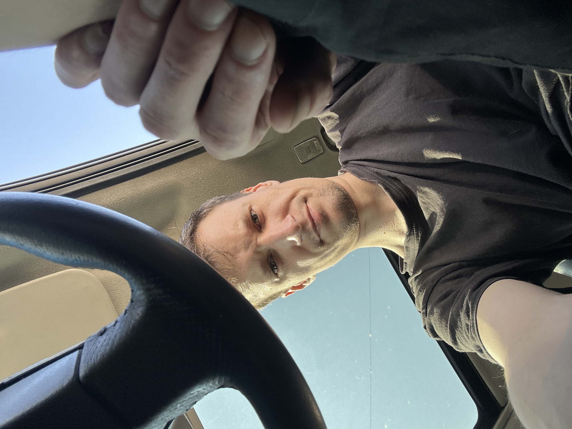 A man with short brown hair and stubble is looking at the camera from inside a car. He is wearing a dark t-shirt. Part of a steering wheel is visible in the upper left corner. A hand is reaching into the frame from the right. The sky is visible through the sunroof of the car, and it appears to be a clear day. The location is not specified as being in Shatava, Ukraine.
