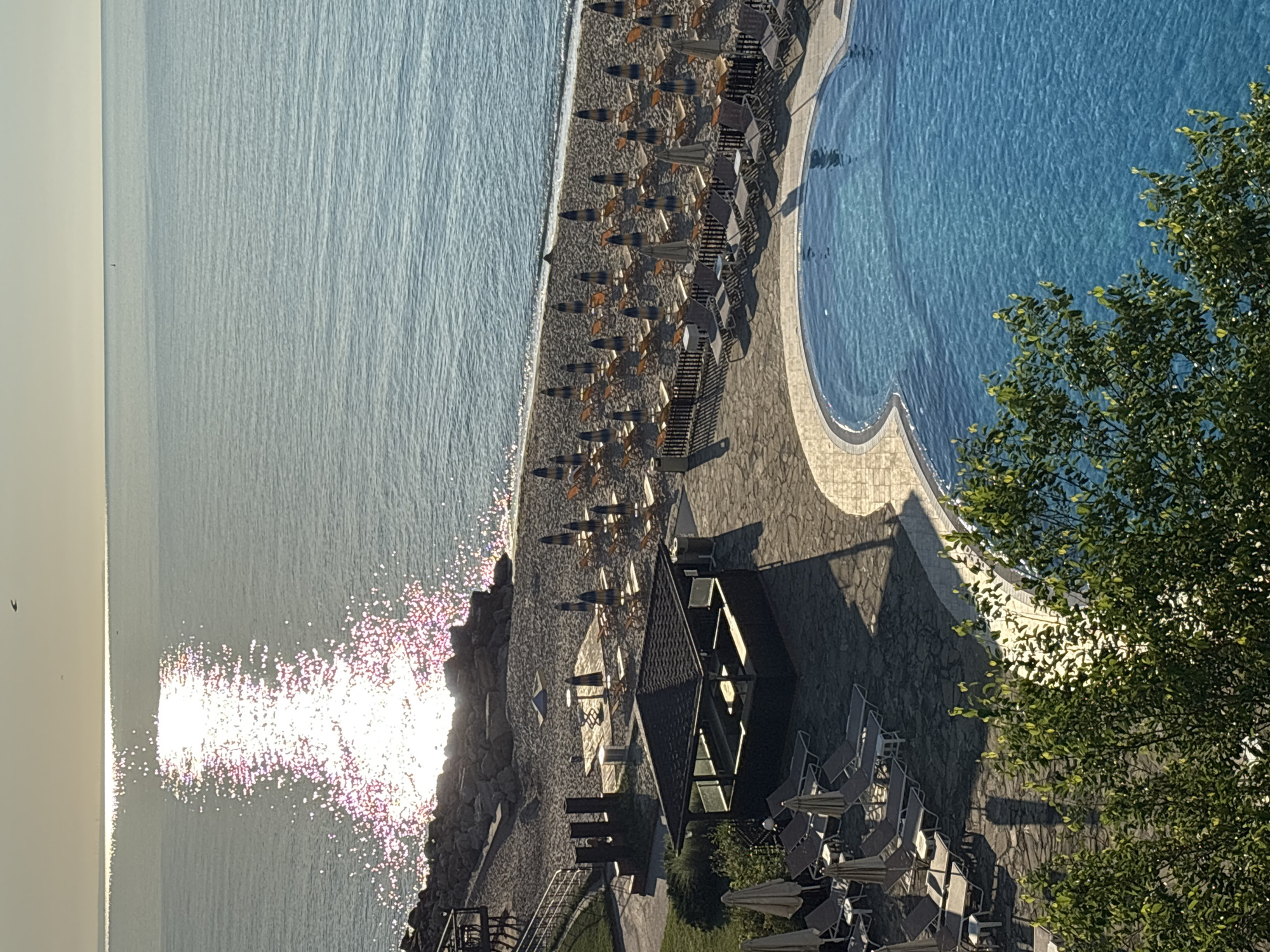 The image shows a sunlit beach and swimming pool area, likely a resort or hotel, overlooking the Black Sea. The sun's reflection creates a glittering path on the water. The beach is lined with numerous closed umbrellas and lounge chairs. A small, dark-roofed open-air structure, possibly a bar or kiosk, is situated on the paved area near the beach. A freeform swimming pool with bright blue water is in the foreground, partially obscured by green foliage. The overall scene suggests a peaceful morning in a coastal resort.