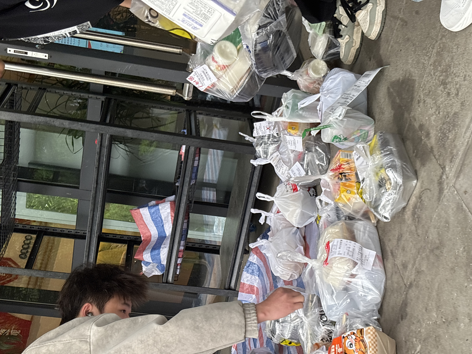 The scene depicts an outdoor or semi-outdoor area in Jiangjin, China, where numerous individually packaged food items are arranged on the ground. A male individual, partially visible on the left, wears a light grey top and has dark hair with an earbud in his left ear. He is leaning forward, with his right hand extended towards several transparent plastic bags resting on a red, white, and blue striped woven tarp. Another individual's lower legs and feet, clad in light-colored sneakers with dark laces, are visible on the far right, standing next to a cluster of packaged items.
The ground is a light grey concrete or paved surface. In the background, a multi-tiered black metal shelving unit is visible, with some shelves empty and one shelf holding a blue and white striped woven plastic sheet. Behind the shelving unit are reflective glass surfaces, showing distorted reflections of greenery and indoor elements. A vertical metal bar, possibly part of a doorframe or support structure, stands to the right of the shelving unit, near the right edge of the frame.
Approximately twenty to thirty various food packages, predominantly in transparent plastic bags, are spread across the ground. Many bags contain white labels with red Chinese characters, likely order or content information. Specific items visible include containers, cups, bread-like items, and other packaged goods, some wrapped in silver foil. The overall arrangement suggests a collection or distribution point for prepared food orders.