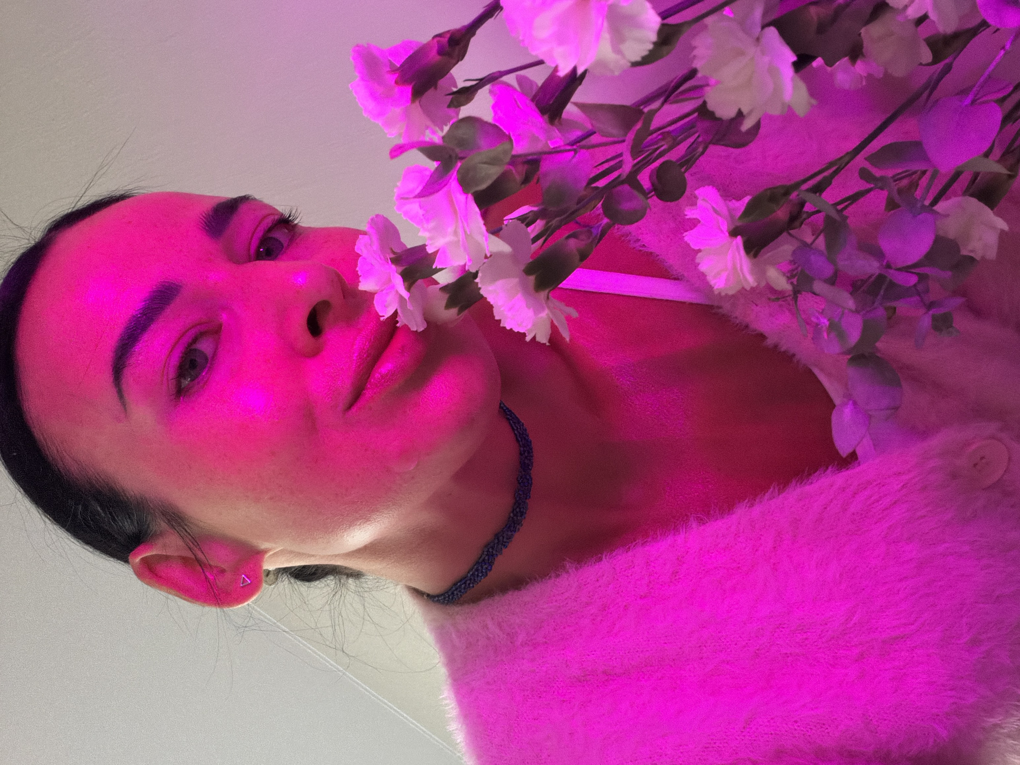 A woman is holding a bouquet of white flowers close to her face. She is wearing a fuzzy, pink sweater and a blue beaded necklace. She has dark hair pulled back, and a small triangular earring in her left ear. The light in the photo is pink, casting a strong magenta hue on the scene. There is no indication of Saint Petersburg, Russia in the image.