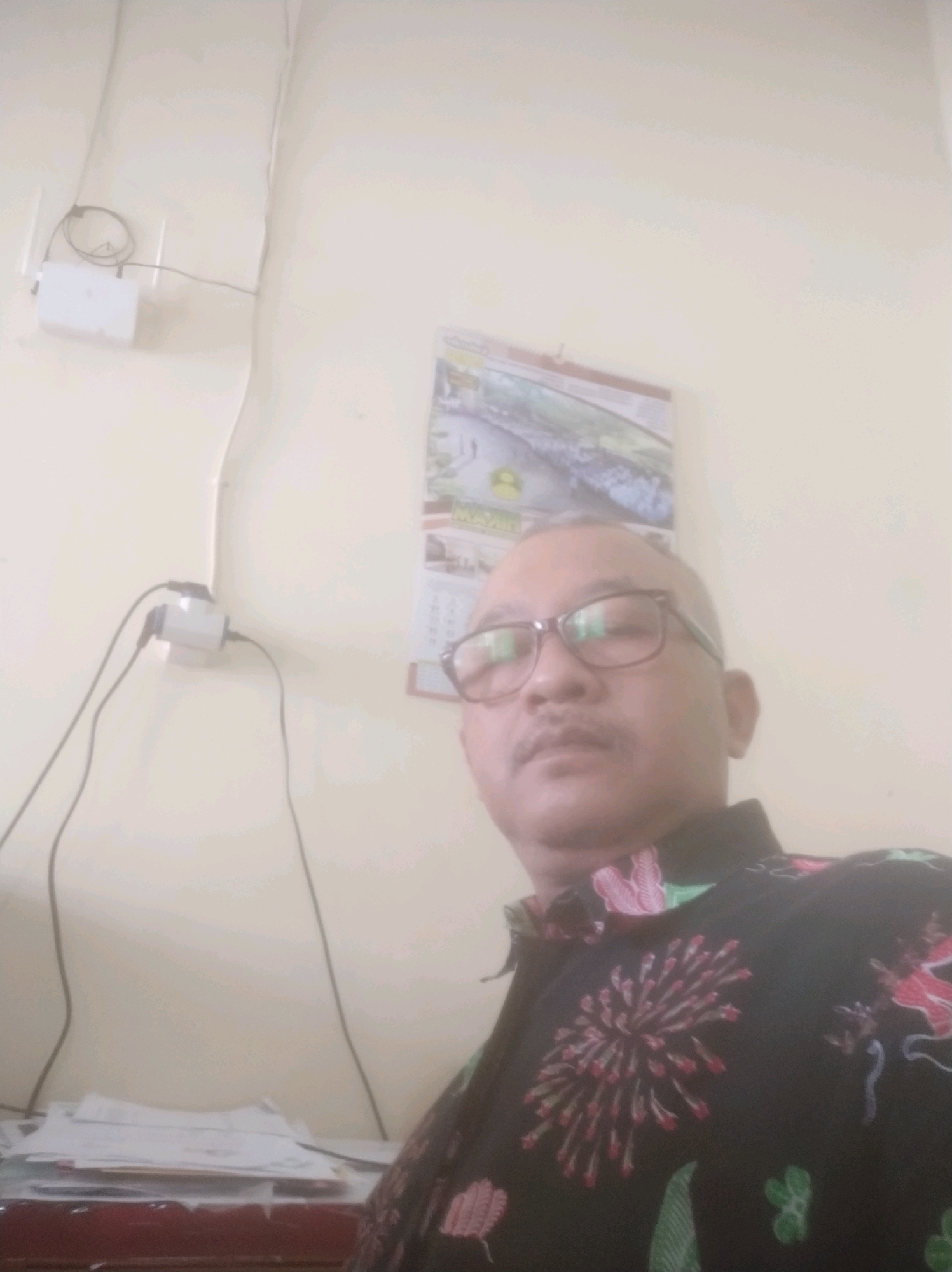 A man with glasses and a mustache is looking at the camera in a room. He is wearing a black shirt with a colorful floral pattern. There is a router mounted on the wall behind him, as well as a calendar hanging on the wall. A stack of papers is visible on a desk in the foreground. The environment appears to be an office or home office. The image does not provide any specific visual cues to determine the exact location in Batan, Indonesia.