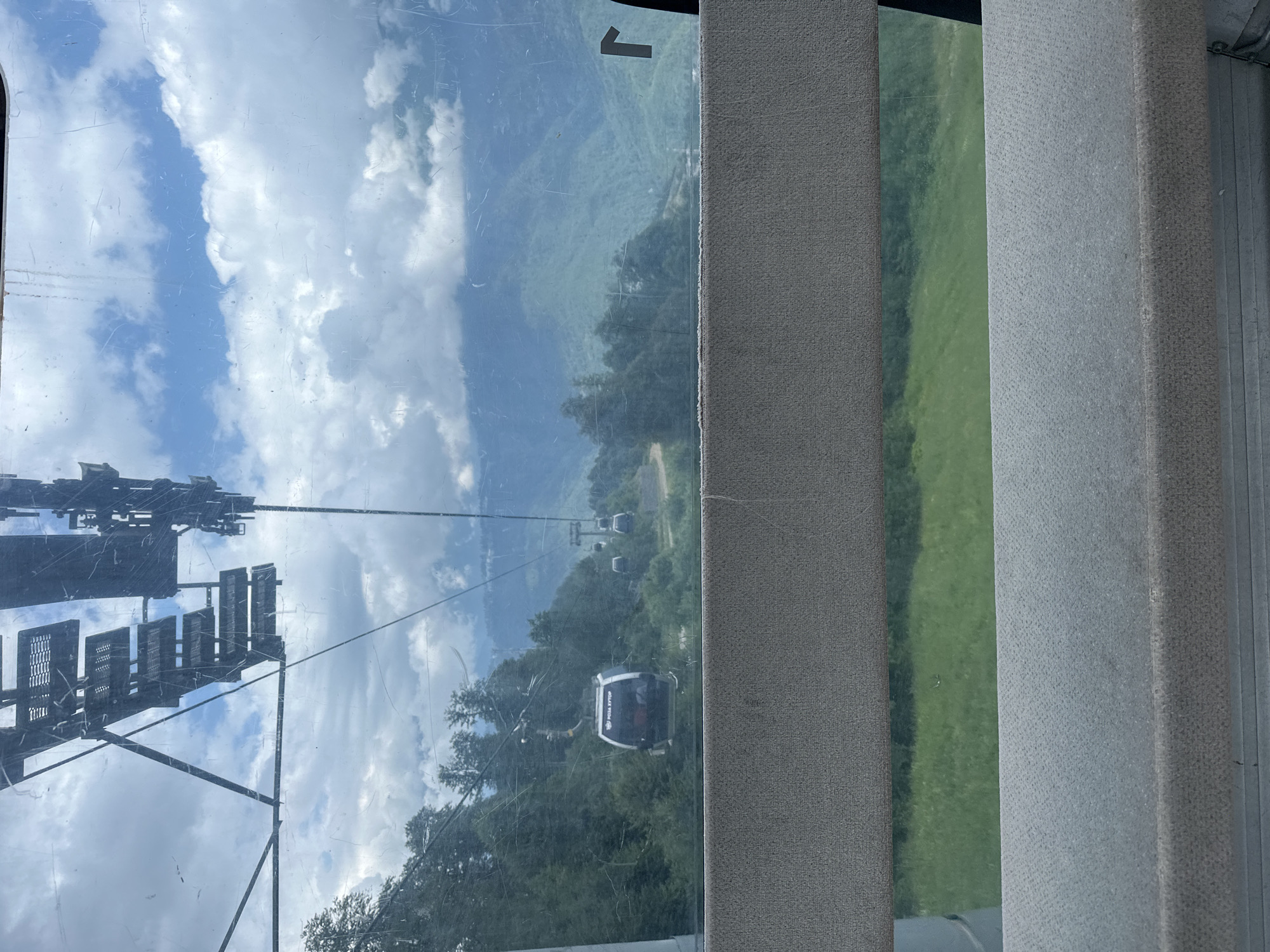 The image depicts a view from inside a cable car, looking out at a mountainous landscape near Estosadok, Russia. The foreground shows the interior of the cable car, with a textured, upholstered barrier running horizontally across the middle of the frame. Through the cable car window, a series of other cable cars are visible, suspended from wires stretching across the frame.
The landscape outside is characterized by lush green forests covering rolling hills and mountains. The sky is partly cloudy, with dramatic white clouds against a blue sky. In the distance, the mountains recede, creating a sense of depth and scale. The cable car in the mid-ground has the Cyrillic text "РОЗА ХУТОР" visible on its exterior. The scene suggests a recreational or tourist setting, typical of a mountain resort.