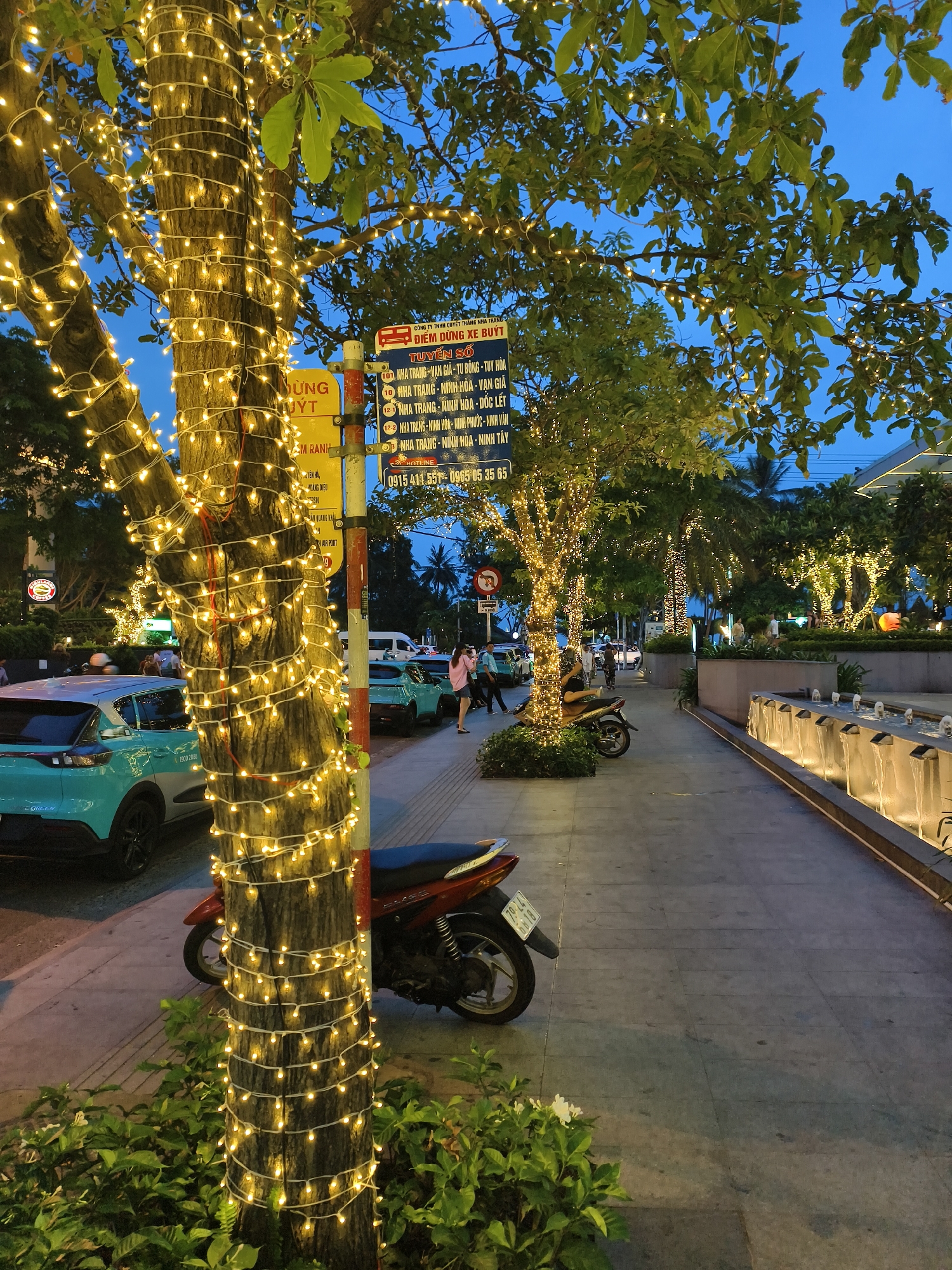 The image shows a nighttime scene on a sidewalk in Nha Trang Ward, Vietnam. Trees are wrapped with strands of yellow lights. On the left is a sign with a yellow background that says "Dừng/Uýt", a stop sign and a blue sign with the numbers of the bus routes in the city of Nha Trang in Vietnamese. A parked red and black motorcycle is in the foreground to the right. Behind the motorcycle is a teal-colored car. Farther down the sidewalk are additional trees also wrapped in lights and a few people walking. On the right is a decorative water feature, and the sky is dark blue.