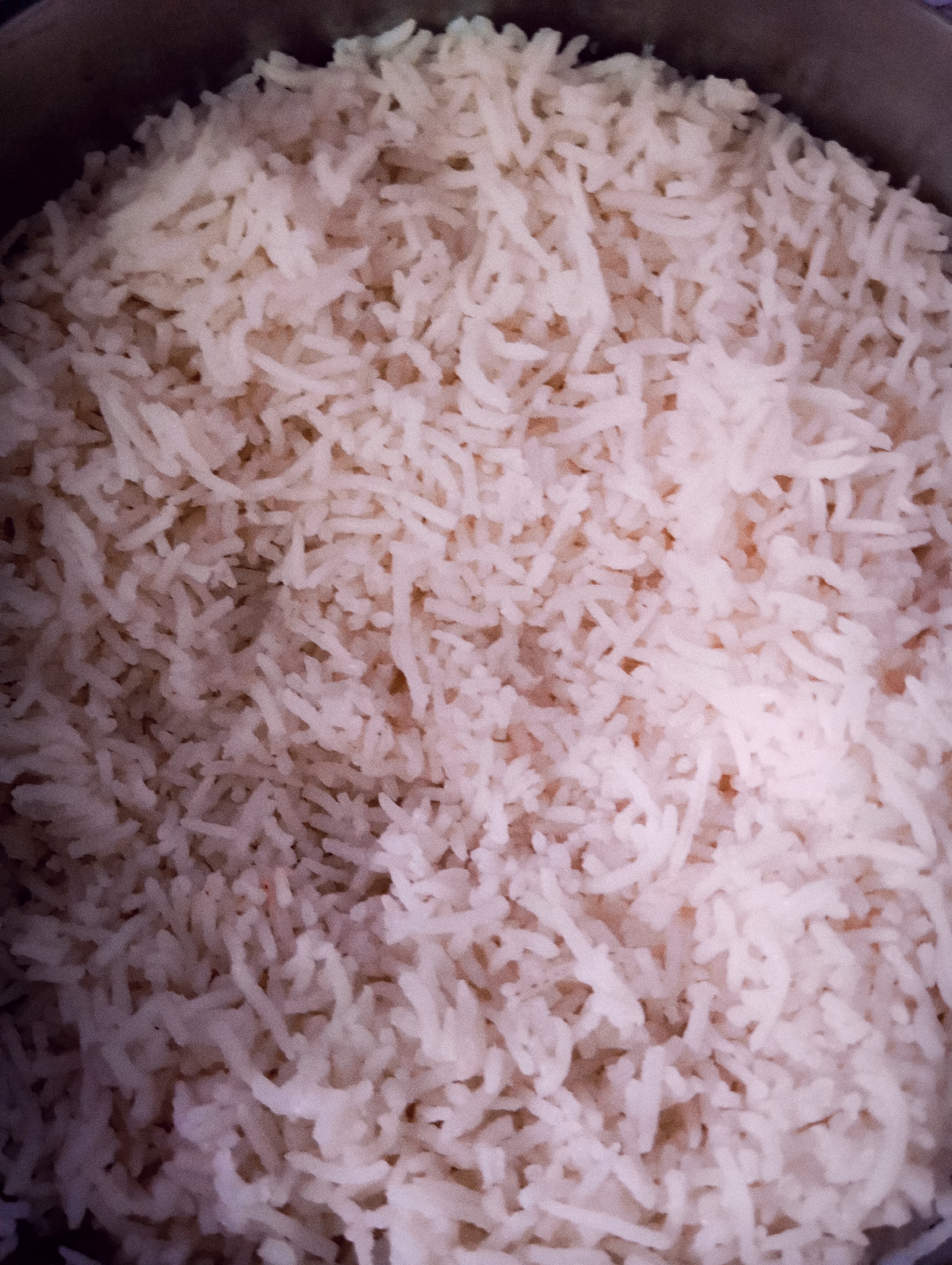 The image is a close-up, overhead shot of a pot filled with cooked white rice. The rice appears to be long-grain and fluffy. The lighting is soft, casting subtle shadows and highlighting the texture of the grains. The edges of a dark pot are visible around the perimeter of the rice. There are no people, animals, or other objects in the frame, and the environment is not discernible. The image is of indeterminate location.