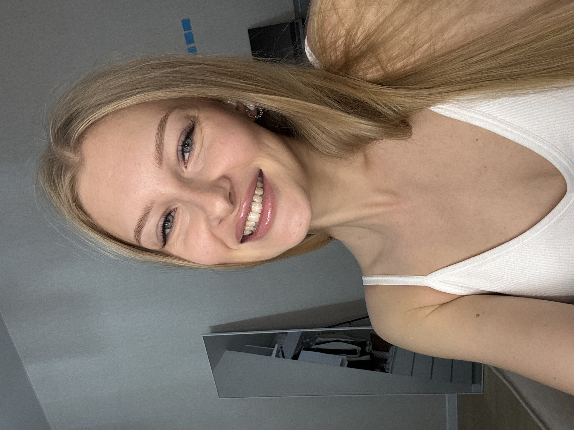 A young woman with long blonde hair and blue eyes smiles at the camera. She is wearing a white tank top and hoop earrings. She is indoors, in what appears to be a bedroom, with a mirror and a closet visible in the background. The location is not specified as Perm, Russia.