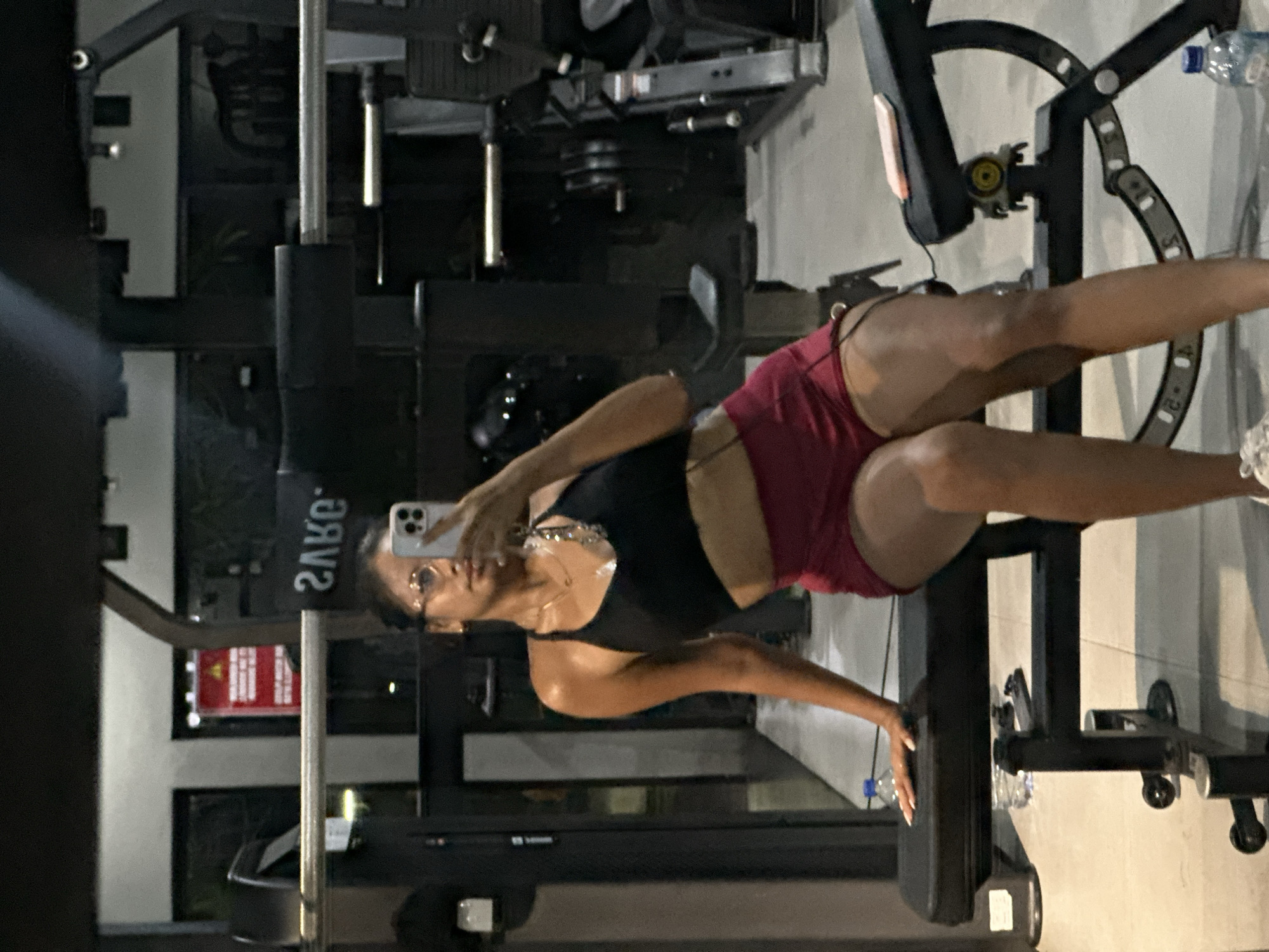 A woman is taking a selfie with her phone in a gym in Batubulan, Indonesia. The woman is wearing a black tank top and red shorts. She is standing in front of a weight machine. There is a water bottle near the weight machine.