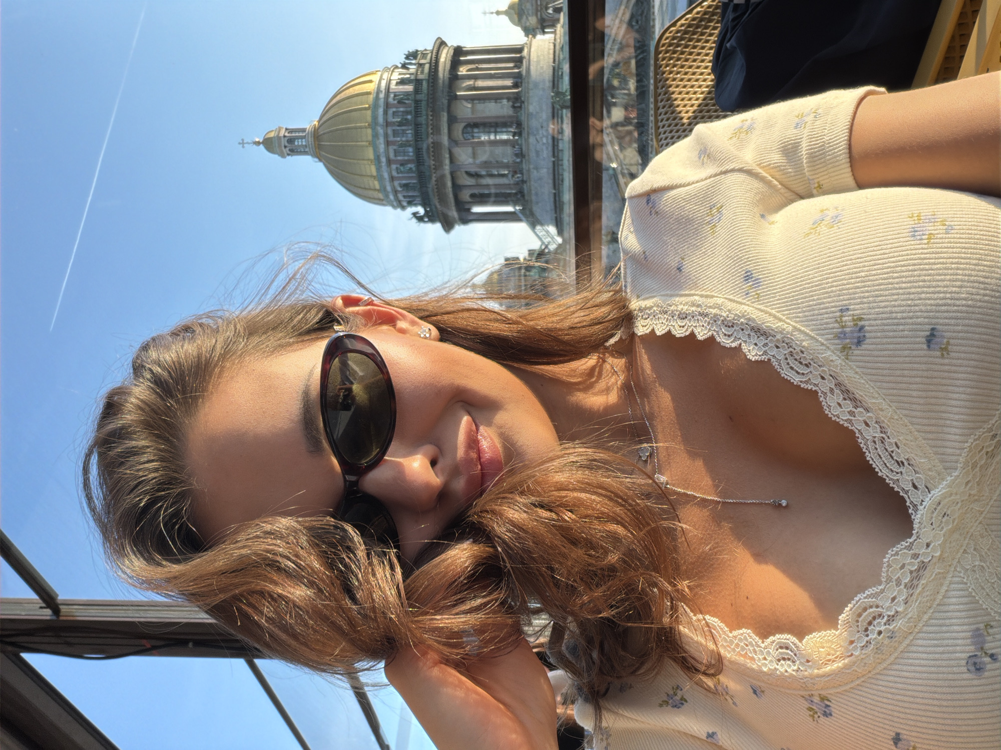 A woman, with medium-length brown hair, is taking a selfie outdoors. She is wearing dark sunglasses, a light-colored top with a floral pattern and lace trim, and a silver necklace with a small pendant. Her left arm is raised, and her hand is touching her hair. In the background, the golden dome of a large, ornate building, identified as St. Isaac's Cathedral in Saint Petersburg, Russia, is visible against a clear blue sky. The sun is casting warm light on the scene.