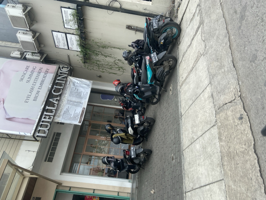 The image shows a street scene in front of a business establishment in Bojongsoang, Indonesia, located at the coordinates -6.972, 107.636. The business is called "Luella Clinio" and advertises skincare, slimming, eyelash extension, and brow embroidery services. Five motorcycles are parked in front of the business, all facing the street. The motorcycles are black, and the front two are a lighter blue. Some motorcycles have license plates visible. The street is paved with concrete, and there is a sidewalk along the front of the building. There is a green vine growing up the wall of the building, and a small amount of greenery in the background. The day is overcast.