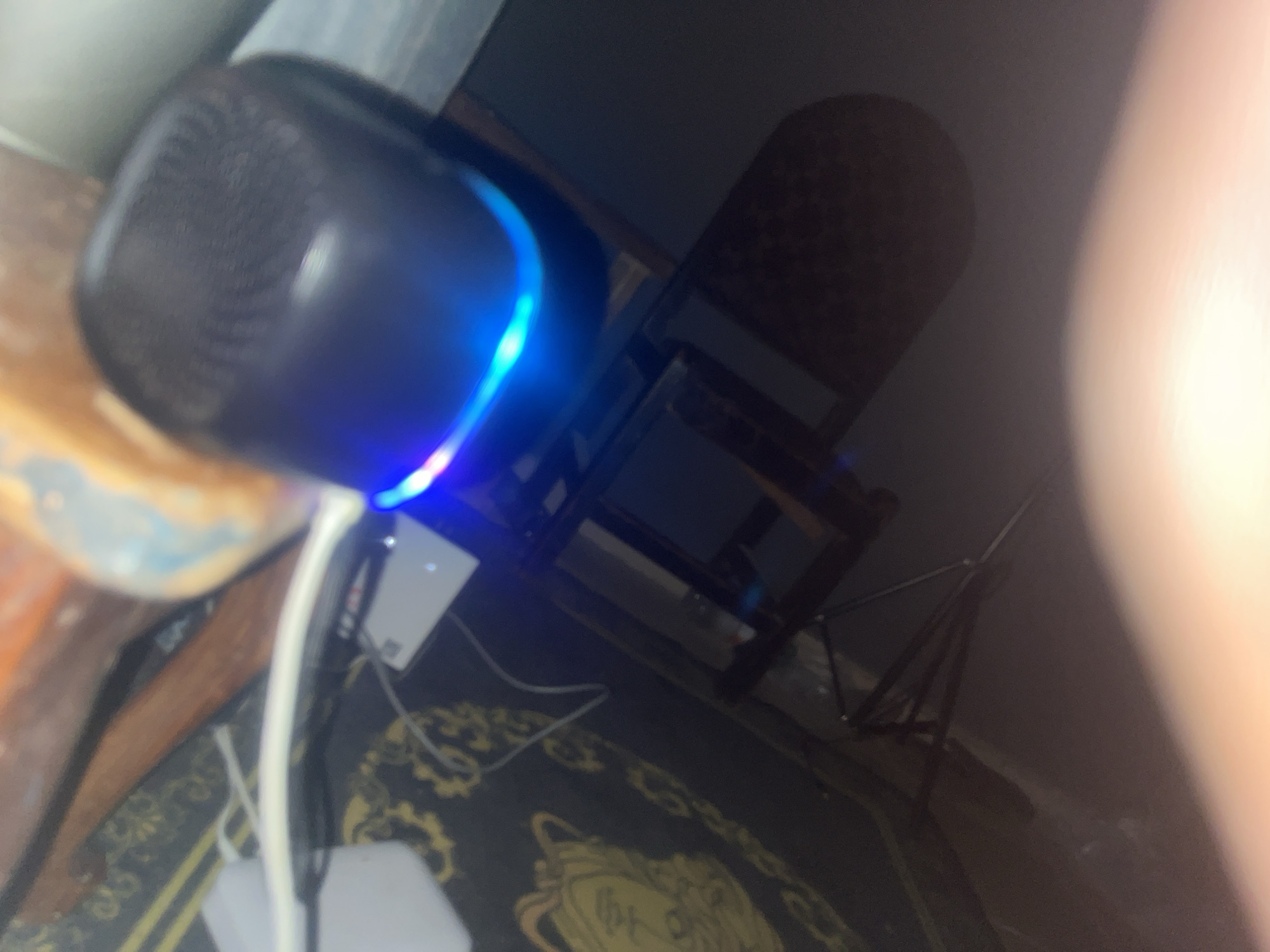 The image shows a close-up of a black Bluetooth speaker with a blue light emitting from its edge. The speaker is sitting on a wooden surface, likely a desk or shelf. A white cable is connected to the speaker. In the background, there is a chair with a dark patterned seat, and a black tripod. The floor has a patterned rug with gold designs. Based on the prompt's request, the image location is in Gura Top, Nigeria. However, the image itself offers no concrete evidence to confirm the location.