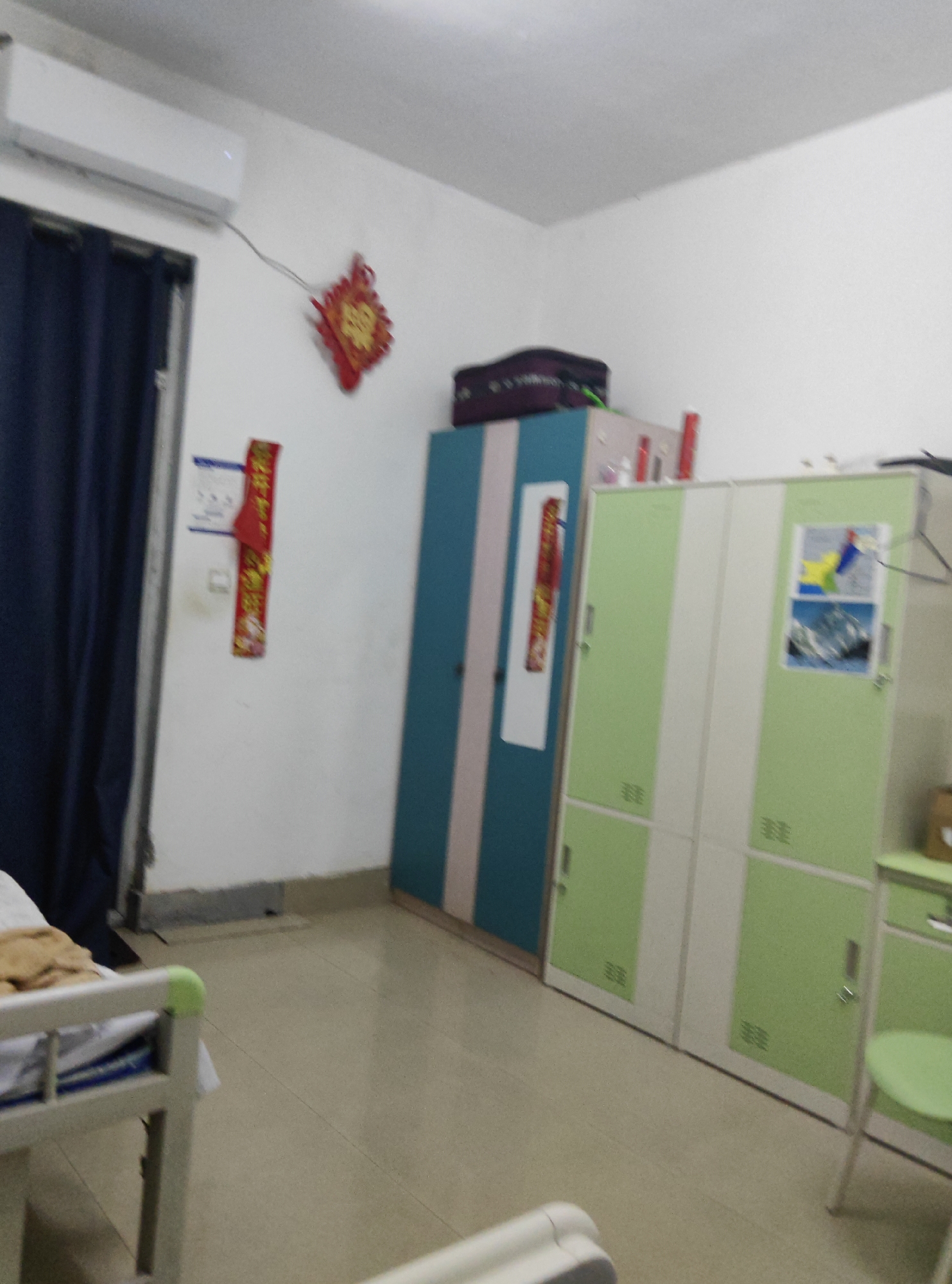 The image depicts a room in a dormitory or hospital, likely located in the Chongqing High-tech Zone, China. The room is sparsely furnished, with a bed frame visible in the foreground. Along the wall, there are several tall cabinets, some in teal and pink stripes, and others in green and white. A dark blue curtain hangs on the left side of the room, partially obscuring an air conditioning unit mounted high on the wall. Decorative items, including red Chinese knots and a long red scroll with yellow characters, are affixed to the wall. A small table with some items on it, including a red bottle, is positioned near the cabinets. The floor is tiled in a neutral color. There are no people visible in the image.