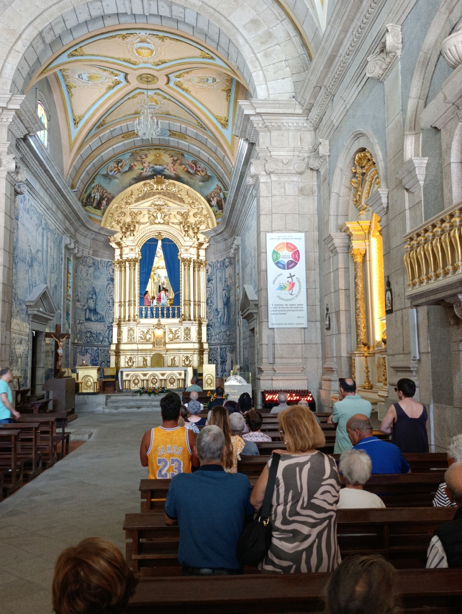 The image depicts the interior of a church in Terras de Bouro, Portugal. The scene is captured from a rear-facing perspective, looking towards the altar at the front of the church. The architecture features high, vaulted ceilings adorned with painted murals and intricate golden decorations. The walls are decorated with large blue and white tiled panels depicting religious scenes.
In the foreground, several individuals are seated on wooden pews, facing the altar. One person in the front row is wearing a yellow basketball jersey with the name "JAMES" and the number "23" on the back. Another person is wearing a black and white patterned dress.
The altar area is highly ornate, with a golden structure surrounding a central display featuring religious figures. Blue curtains are drawn behind the figures. A large, decorative crucifix is visible to the left of the altar.
A banner with colorful circular graphics and text in Portuguese hangs on a pillar to the right of the altar. The lighting in the church appears to be a combination of natural light coming from windows and artificial lights. The overall atmosphere suggests a religious service or a visit to a place of worship.