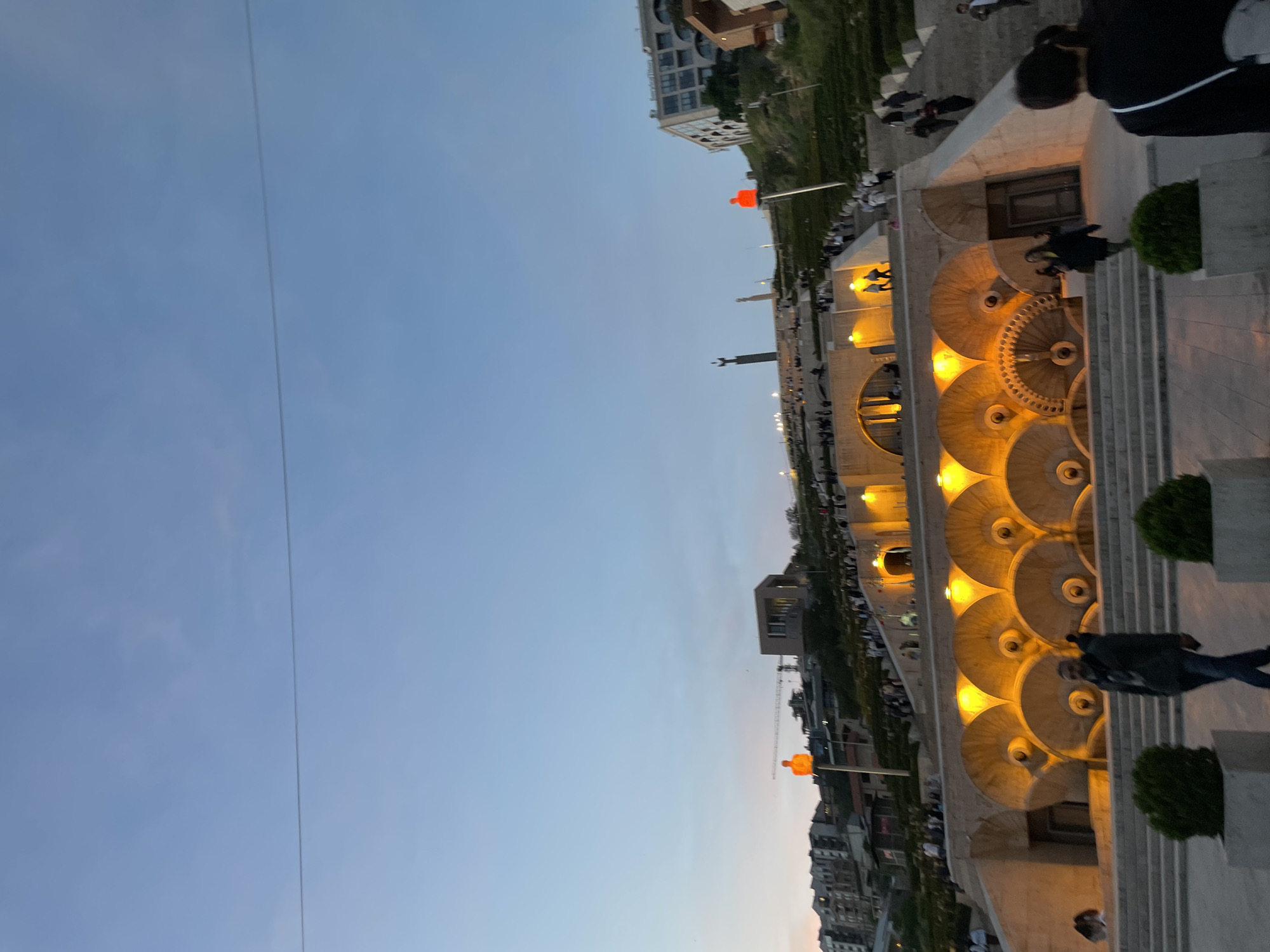The image shows the Cascade in Yerevan, Armenia, a monumental stairway with decorative water features. People are seen walking along the steps. The architecture is illuminated, contrasting with the dusk sky. In the background, urban buildings and an orange sculpture are visible. A utility cable is visible in the sky.