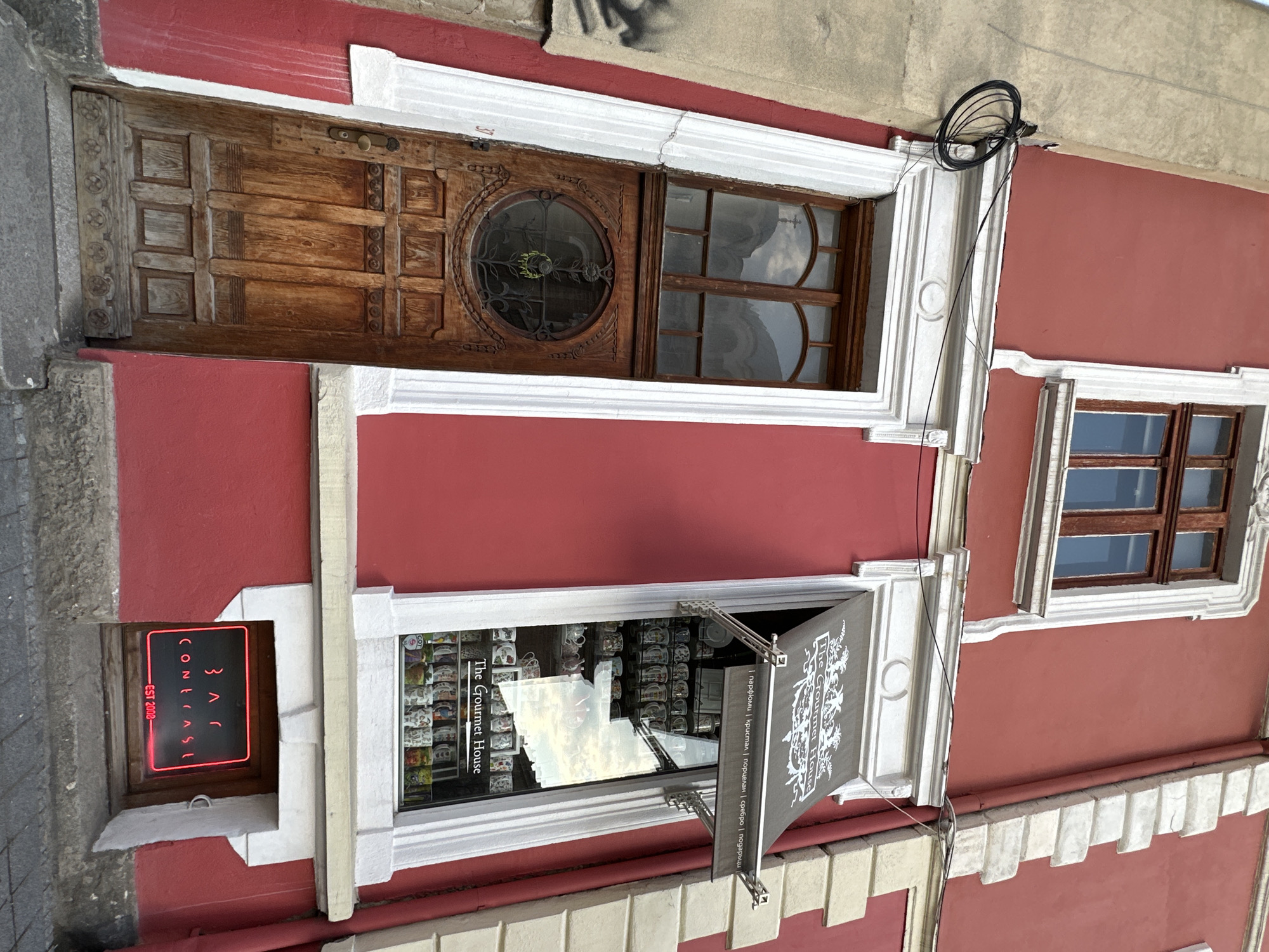 The image is of a red building, likely a shop, located in Sofia, Bulgaria. The building has four windows and a door. The top window is next to a wooden door with an oval window. The next window down has a sign that reads "The Gourmet House" and has a banner hanging from it with Cyrillic text. The bottom left window has a neon sign that reads "Bar Contrast Est 2008". The final window on the right has small windows. The building has white trim. There are wires hanging from the top of the building.