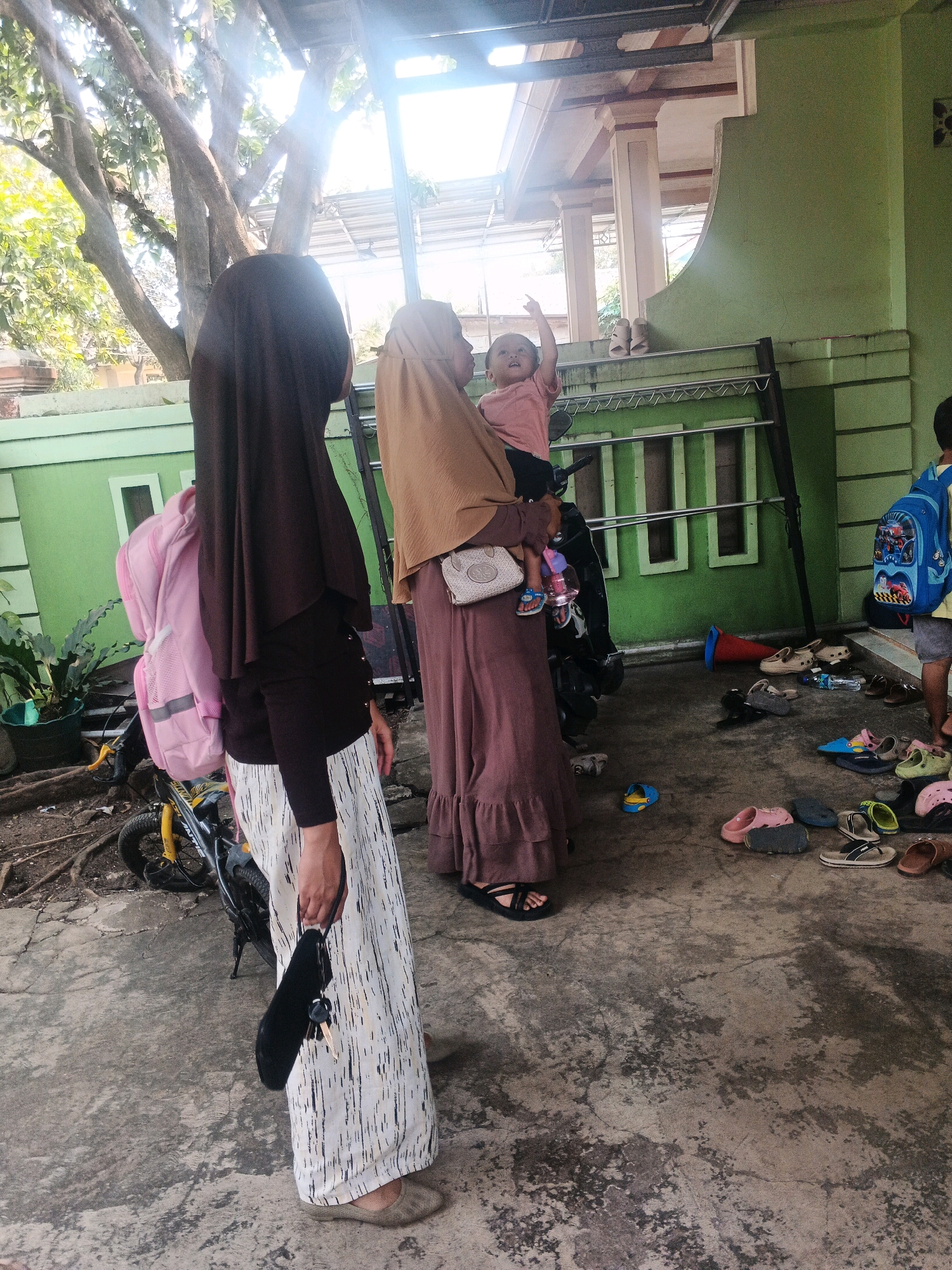 In Kadu, Indonesia, a group of people are gathered outdoors on a concrete surface in front of a green building. A woman wearing a dark brown hijab and a white and black patterned skirt stands on the left, holding a black clutch with keys. To her right, another woman in a light brown hijab and a long, rust-colored dress holds a baby who is pointing upwards. Behind them, a child wearing a blue backpack stands near the green building. Various pairs of shoes and sandals are scattered on the ground around them. A bicycle with a pink backpack is parked to the left. The environment appears to be a residential area with trees and a partially visible covered structure overhead.