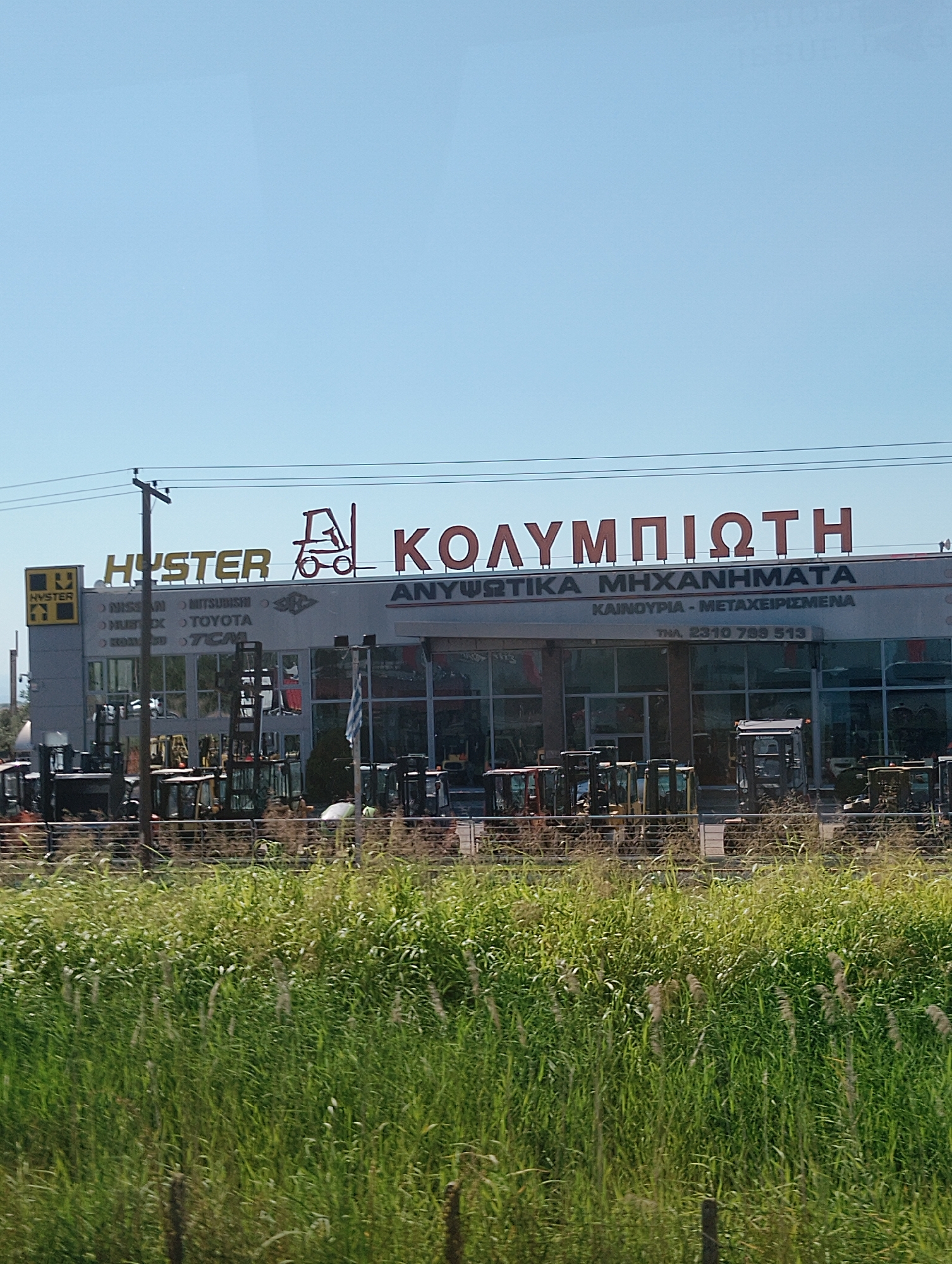 The image shows the facade of a commercial building. The building has a large sign with red lettering that reads "ΚΟΛΥΜΠΙΩΤΗ" and "ΑΝΥΨΩΤΙΚΑ ΜΗΧΑΝΗΜΑΤΑ" above a smaller sign that reads "ΚΑΙΝΟΥΡΓΙΑ - ΜΕΤΑΧΕΙΡΙΣΜΕΝΑ" and a phone number "ΤΗΛ. 2310 788 513". Another sign on the left reads "HYSTER" with a yellow logo. Below the signs, a row of industrial vehicles, likely forklifts and other heavy machinery, are parked outside the building. The scene is viewed from across a field of tall, green grass. The sky above is clear and blue. Based on the text on the building, this appears to be a dealership or service center for lifting machinery and vehicles, potentially located in the Municipal Unit of Echedoros, Greece.