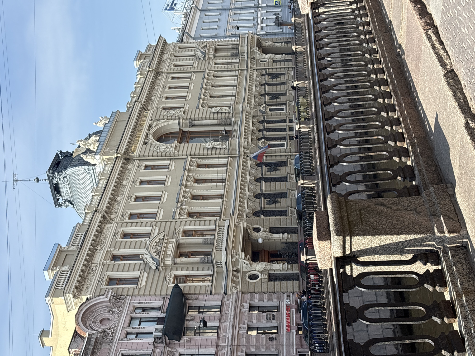 An eye-level medium shot depicts a section of the ornate, beige-colored building at 24 Nevsky Prospect in Saint Petersburg, Russia. The building facade is highly detailed with architectural elements, including numerous windows, decorative moldings, and sculptural details. A Russian flag is visible, protruding from the side. Adjacent to the building is a bridge. Ornate ironwork railings run along the bridge's edge. Several cars are parked along the street below. The sky above the building is blue.