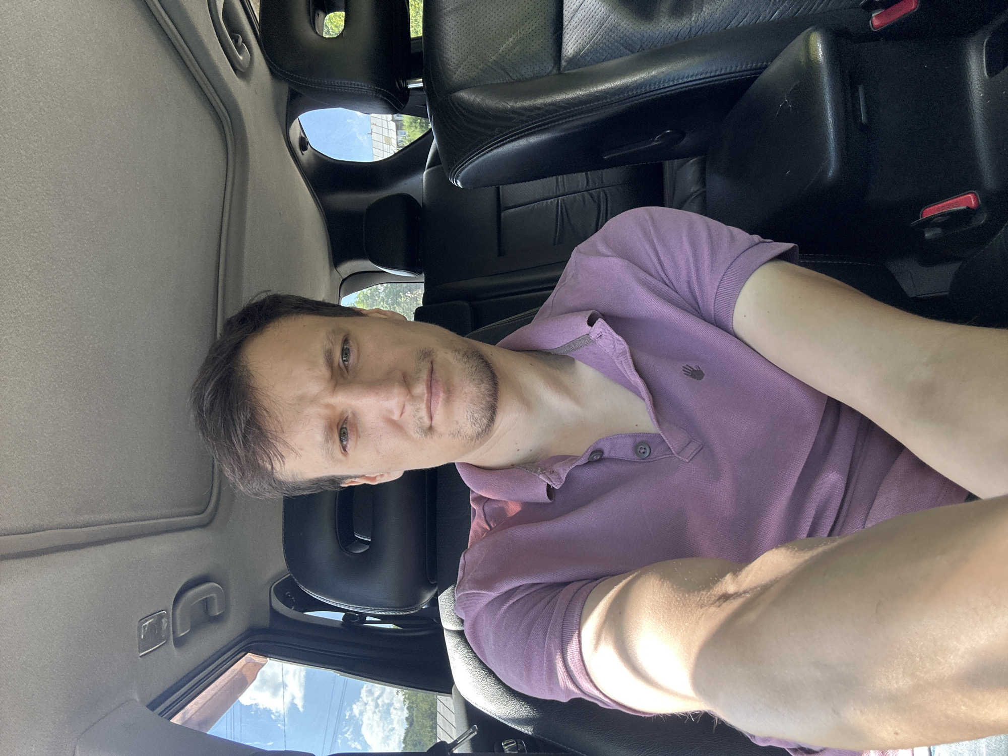 The image shows a man sitting in the driver's seat of a car, taken from a selfie perspective. He is wearing a purple polo shirt. The car's interior is visible, including the headrests of the passenger seats and a sunroof. Sunlight is streaming in, casting shadows on the man's arm and torso. The background visible through the car windows suggests an outdoor setting, possibly with greenery and a building. The man appears to be in his late 20s or early 30s, with short brown hair and a light beard. He is looking directly at the camera with a neutral expression. There is no indication that this scene is located in the city of Makiv, Ukraine.