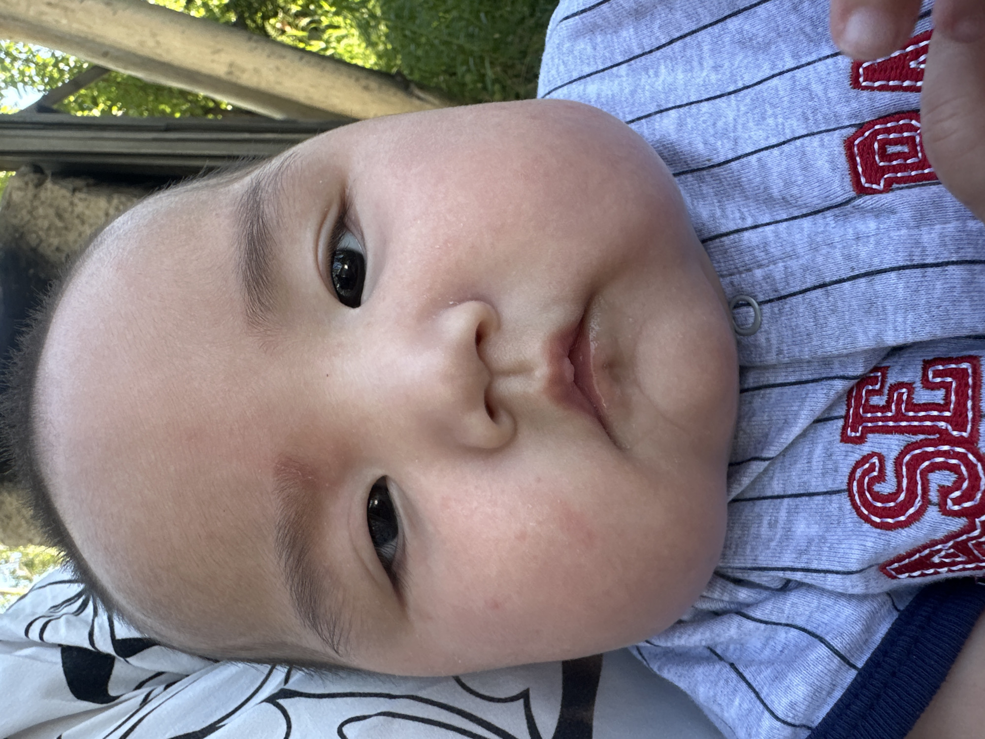 The image shows a close-up, low-angle shot of a baby's face. The baby is lying down, with its head tilted upwards. The baby has dark eyes and dark hair, and its cheeks are round and rosy. The baby is wearing a gray and white striped shirt with red lettering. The background is blurred, but appears to be outdoors, with green foliage and wooden structures visible. The lighting suggests it is daytime, possibly sunny. The baby's expression is neutral, with its mouth slightly open. There is no discernible activity or event taking place, other than the baby possibly being held or positioned for the photograph. The location context provided is "Тайгараев, Kyrgyzstan", but there are no specific visual cues in the image to confirm this location. The red lettering on the shirt is partially visible but the full text is not legible.
