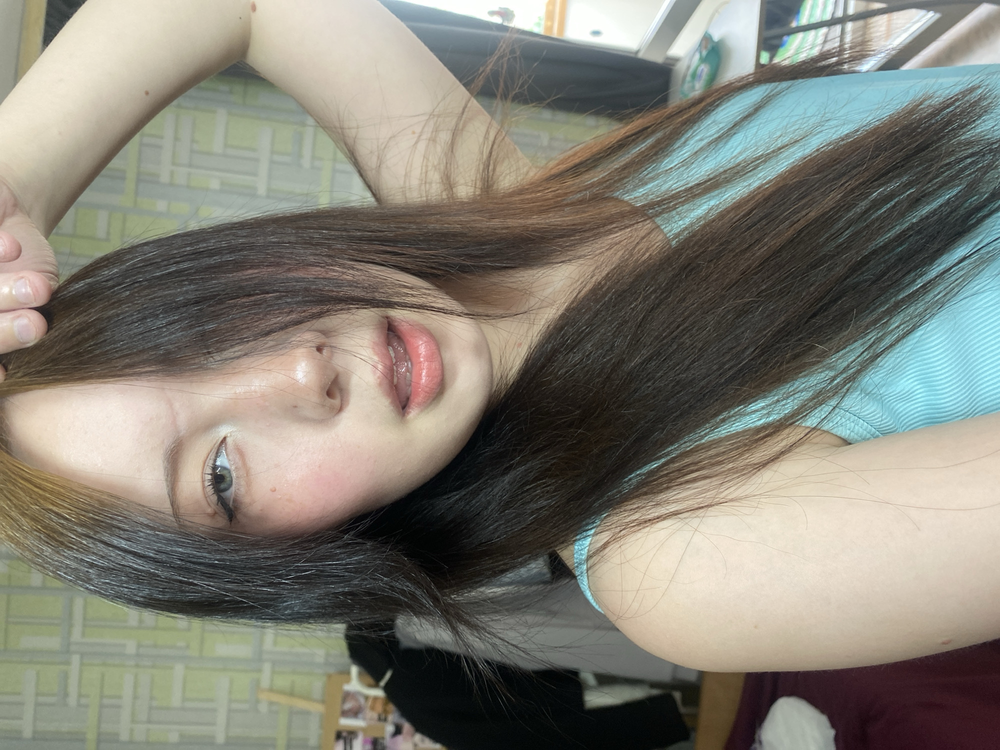 A close-up, selfie-style, rotated image of a young person with fair skin and long, dark brown hair with lighter highlights on the left side. The person is wearing a light blue, striped tank top. Their right arm is raised above their head, with the hand visible in the upper left corner. The person has a light eye color. A portion of a wall with a grid pattern can be seen in the background. Behind and below the person, there are indistinct shapes, possibly of clothing or furniture. The location and city are unknown.