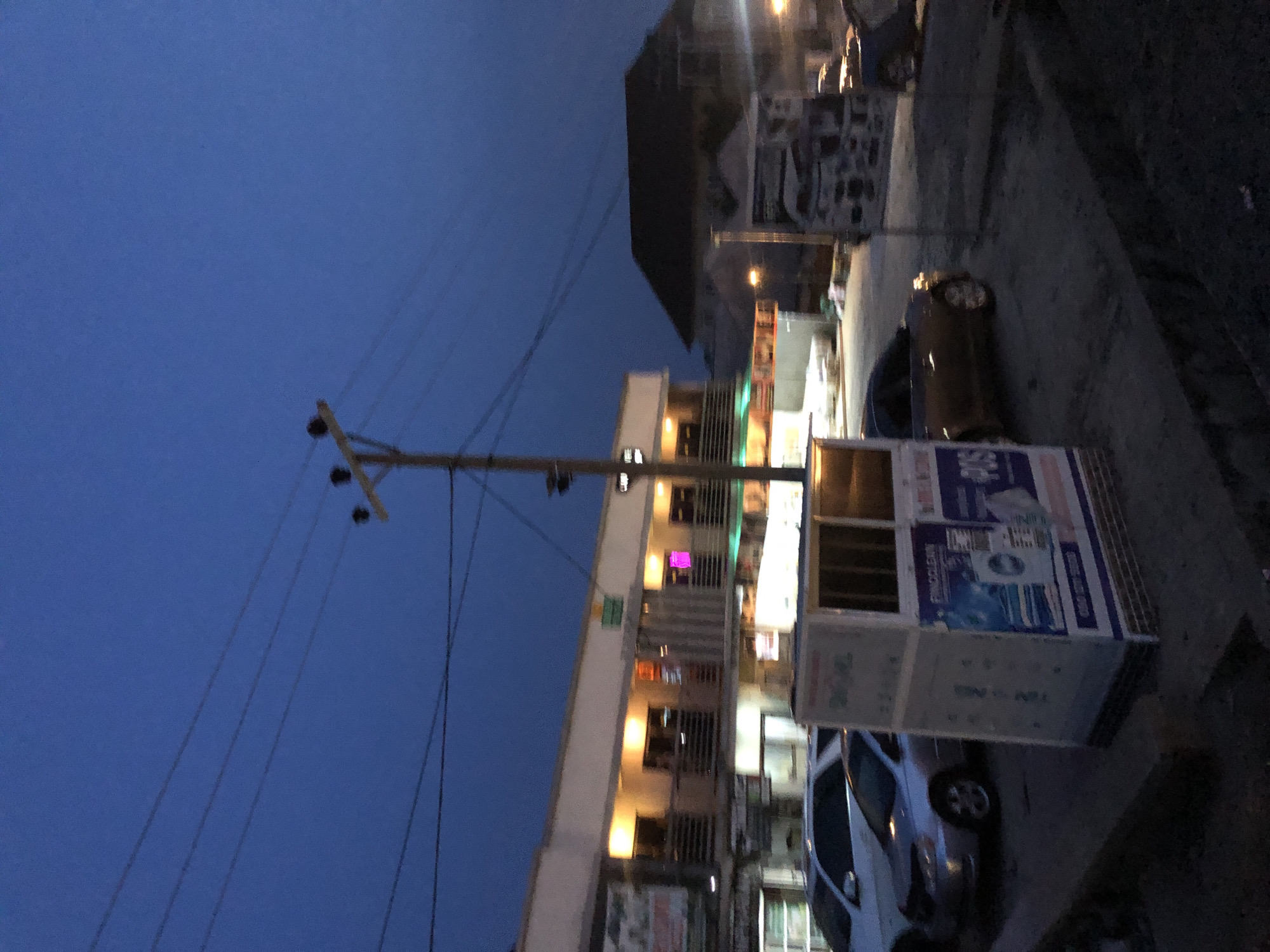 The image captures a street scene in Karu, Nigeria, during dusk or nighttime, characterized by a deep blue sky transitioning from day. A prominent wooden utility pole with numerous power lines extends from the left, reaching towards the upper frame.
Multi-story buildings line the right side of the street, their windows and some storefronts illuminated with artificial light, casting warm yellow, white, and a distinct pink glow from different levels, indicating ongoing activity. In the foreground, a white and blue kiosk-like structure stands on the sidewalk. This kiosk features visible text and graphics, including "FINGOLEXX," "POS" (likely indicating Point of Sale services), and partial words such as "TM NE" and "TIN NING." It also displays imagery resembling an appliance, possibly a washing machine, accompanied by graphics that suggest water or cleanliness.
Several vehicles are present on the street; a dark-colored car is partially visible further down the street, and a lighter-colored, possibly silver, sedan is parked near the kiosk. The ground appears to be paved. The scene portrays an active urban or semi-urban environment after sunset, with artificial lights providing illumination. No individuals are clearly visible in the frame.