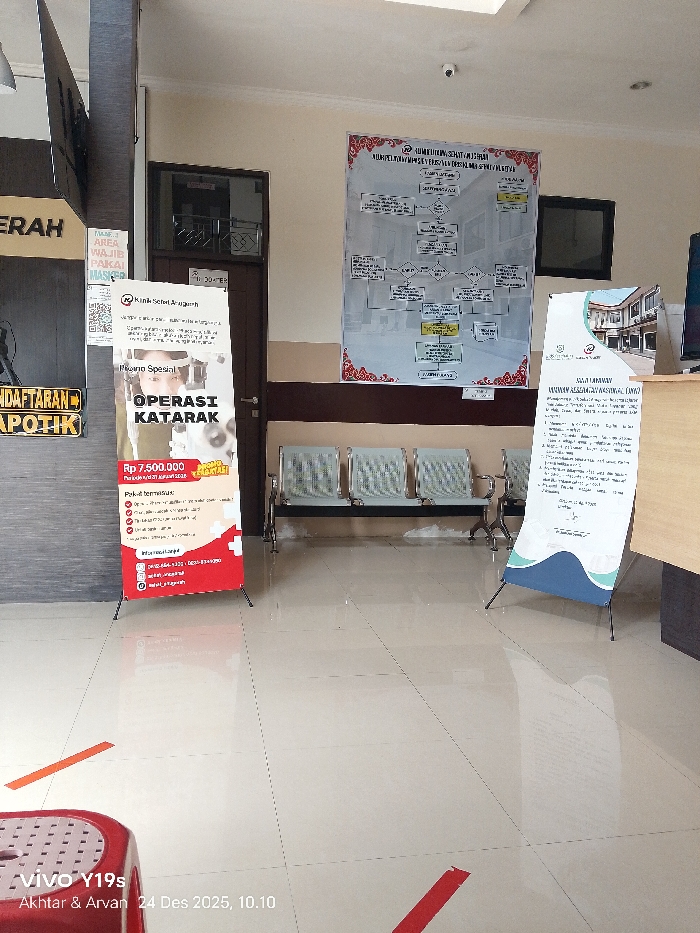 A waiting area inside a building, possibly a clinic or hospital. There is signage indicating "ERAH AREA WAJIB PAKAI MASKE" suggesting mask requirements, "PENDAFTARAN APOTIK," and a banner advertising "OPERASI KATARAK" (cataract surgery) for Rp 7.500.000. There are chairs in the waiting area, information posters on the wall, and floor markings for social distancing. The watermark "Vivo Y19s" and a date are visible in the bottom corner.