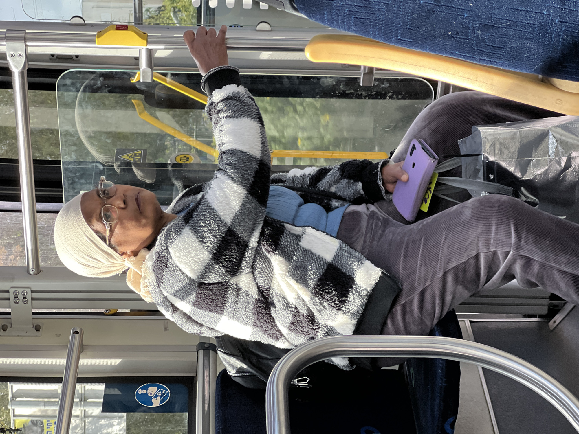 An elderly woman with dark skin, wearing a white headscarf and glasses, is seated on a bus in Kiryat Motzkin, Israel. She is wearing a blue top and a black and white checked jacket. She is also wearing grey corduroy pants. She is holding onto a railing with her right hand and holding a purple phone in her left hand. A shiny silver shopping bag is in her lap. The interior of the bus is visible, including the windows, railings, and other seats.