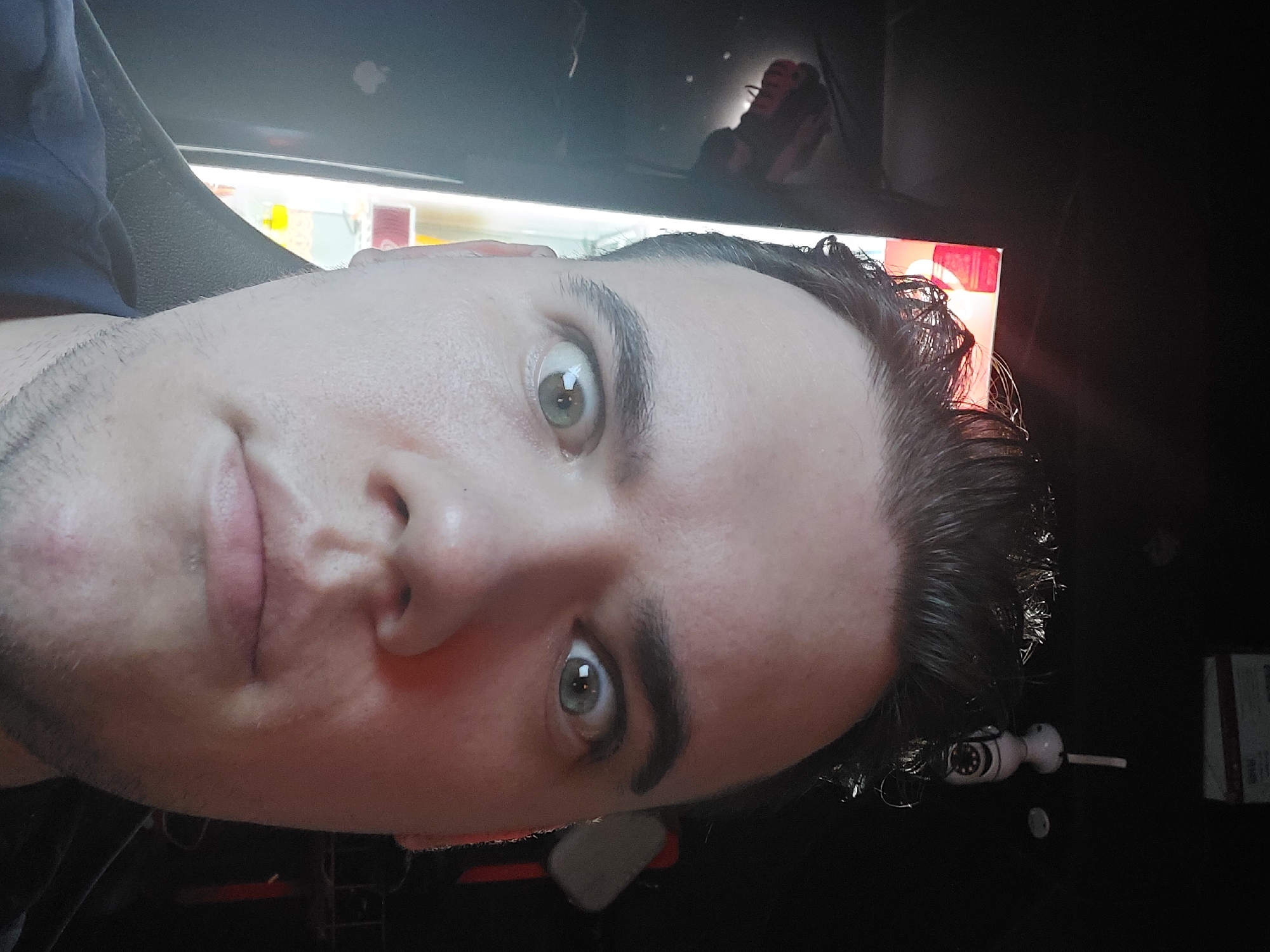 A close-up, rotated image shows a man's face with fair skin, short dark hair, and light-colored eyes looking at the camera. The background includes a vending machine with illuminated products and dark surroundings. There is a white and black object near the right edge of the frame. No evidence in the image suggests that the location is Tashkent, Uzbekistan.