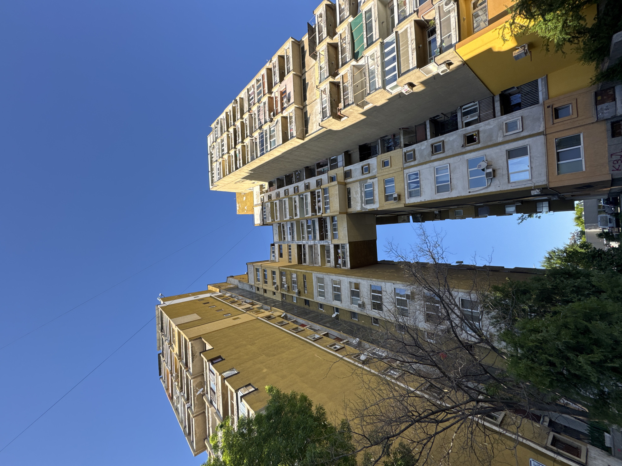 The image shows a unique architectural structure in Sofia, Bulgaria, commonly known as the "Vladislavova House." Two tall, slender buildings stand close to each other with an elevated connection spanning between them, creating a bridge-like structure high above the ground. The buildings have a utilitarian, modernist design with multiple windows of varying shapes and sizes. The color scheme is predominantly yellowish-beige, with some sections in white and gray. The sky is clear and blue, providing a stark contrast to the buildings' earthy tones. Part of a tree with bare branches and green foliage is visible in the foreground, further emphasizing the urban setting. Overhead wires extend across the sky in the left portion of the frame.