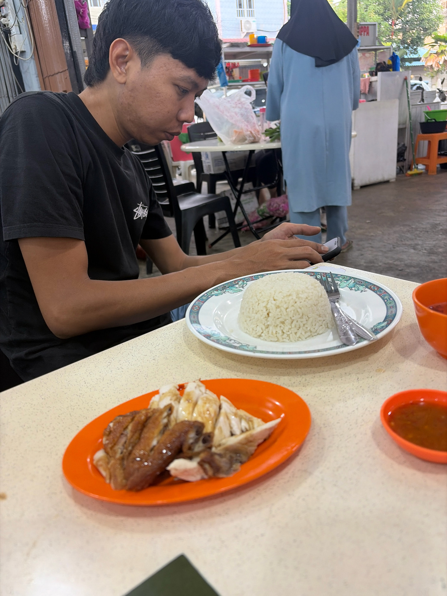 A man, likely of Southeast Asian descent, is seated at a table in what appears to be an outdoor food stall or hawker center in Tanjung Pinang, Indonesia. He is wearing a black t-shirt and is looking down at his mobile phone. In front of him are two plates of food. The plate on the right contains a mound of white rice with a fork and knife. The plate on the left, an orange plate, holds pieces of roasted chicken and what seems to be chicken broth. To the right of these plates is a small orange bowl containing a reddish-brown sauce, likely a dipping sauce for the chicken. In the background, a woman in a light blue hijab and matching outfit stands near a white refrigerator. The environment suggests a casual dining setting with tables, chairs, and other food stalls visible in the distance. The overall atmosphere is that of a typical street-side eatery.