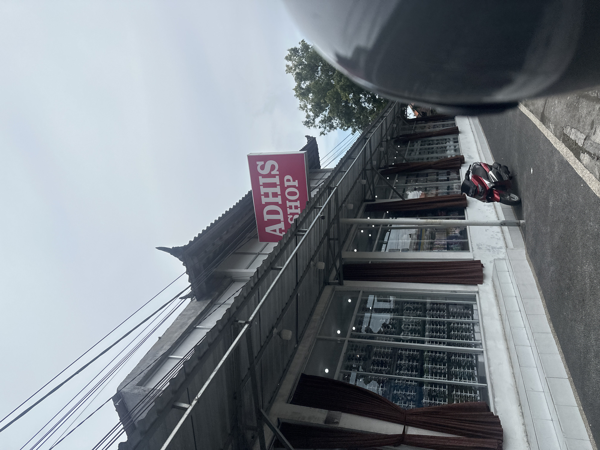 The image depicts a commercial building identified as "ADHI'S SHOP" in Batubulan, Indonesia, viewed from a street-level perspective under an overcast sky. The shop is a single-story structure with a white facade and a prominent red sign displaying "ADHI'S SHOP" in white lettering. The building features multiple large glass windows along its front, behind which dark brown curtains are drawn partially. The windows display numerous items, consistent with optical goods or spectacles, arranged on white shelving units. An extended awning, supported by a visible metal framework, covers the sidewalk area in front of the shop. A decorative, dark-tiled roof element, indicative of traditional Balinese architecture, is visible on the building's right side. Multiple black utility wires traverse the upper left quadrant of the sky. A red and black scooter, identified as a Yamaha NMAX, is parked facing left on the asphalt pavement directly in front of the shop. A portion of a dark grey motorcycle helmet is visible in the lower right foreground, partially obscuring the scene. Green tree foliage is visible above the shop's roofline on the far right. No individuals are present in the observable scene.