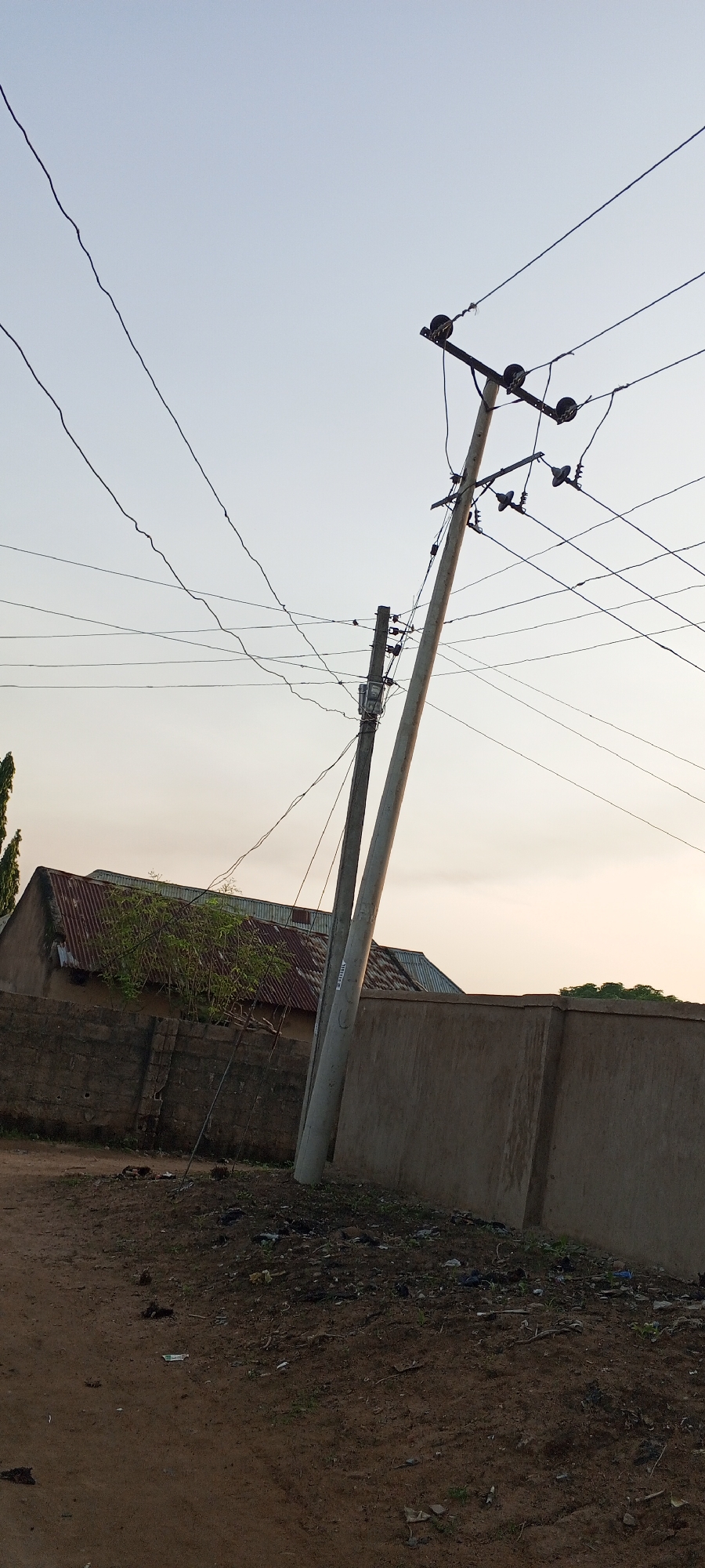 A medium shot captures a street scene in Jalingo, Nigeria, dominated by a leaning utility pole adorned with numerous electrical wires. Another, more slender utility pole stands to its left, also bearing wires and what appears to be a meter. The ground in the foreground is a mix of dirt and debris, with some scattered litter. Behind the utility poles, a low, rough-textured concrete wall runs horizontally across the frame. Beyond the wall, the corrugated metal roof of a building is visible, with some greenery encroaching on it. The sky above is a pale, hazy blue, suggesting either early morning or late afternoon light.
