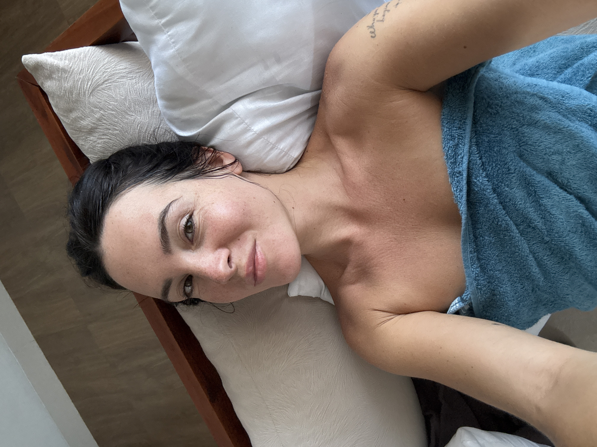 A woman lies on a bed in North Kuta, Indonesia, wrapped in a blue towel. Her hair is dark and pulled back. She is looking at the camera and smiling slightly. There are pillows behind her and a wooden headboard to her left. The floor appears to be tiled with a wood-like pattern.