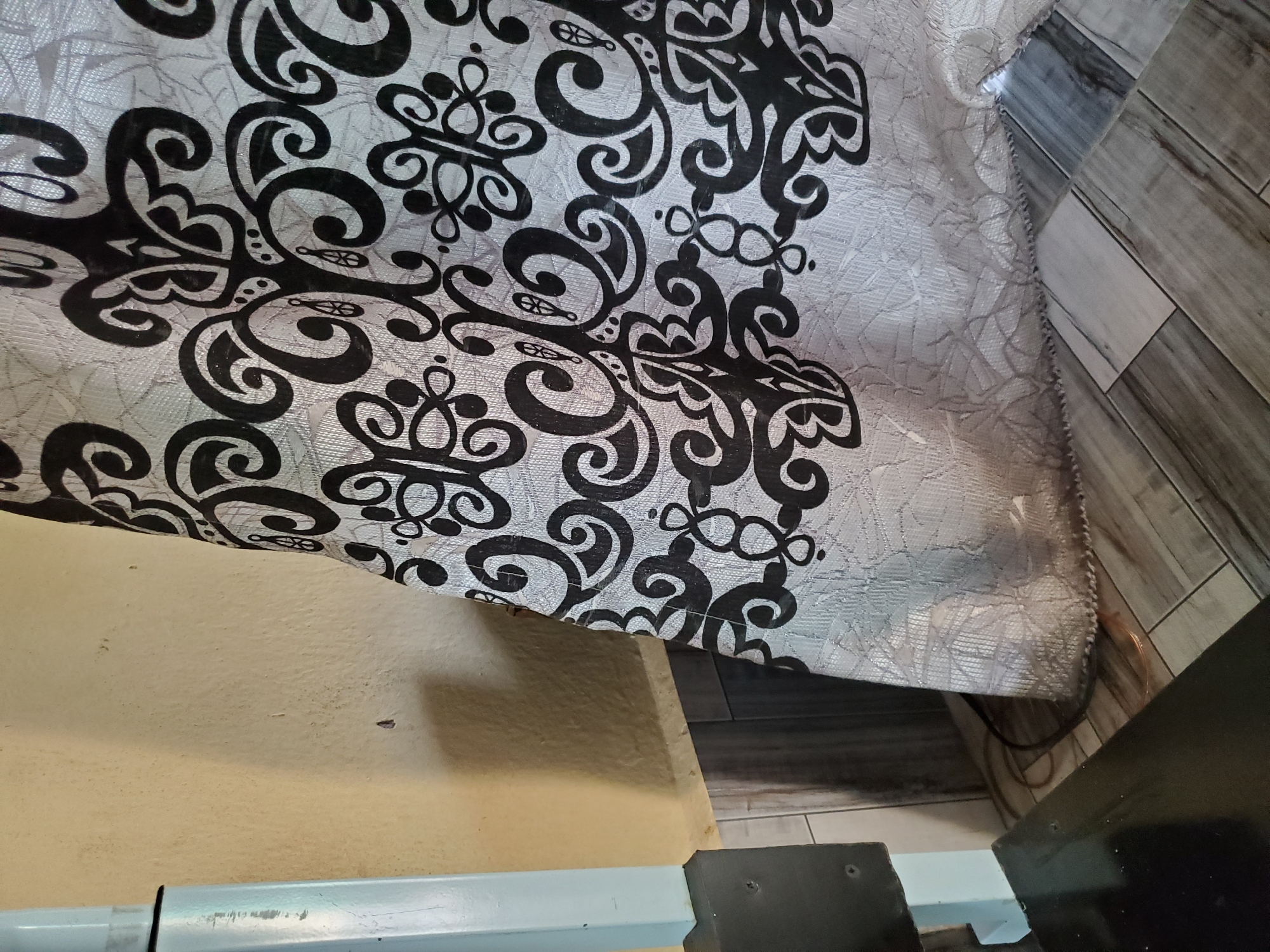 The image shows a close-up of a patterned curtain with black and white swirls hanging in front of a textured yellow wall and a dark-colored floor with a wood-grain pattern. To the left of the curtain, a light blue metal object, possibly part of a machine or piece of furniture, is visible. The curtain's hem rests on the floor, near a coiled brown electrical cord. The scene appears to be an indoor setting, likely a room in Jalingo, Nigeria, given the context provided. There are no people or actions depicted in the image.