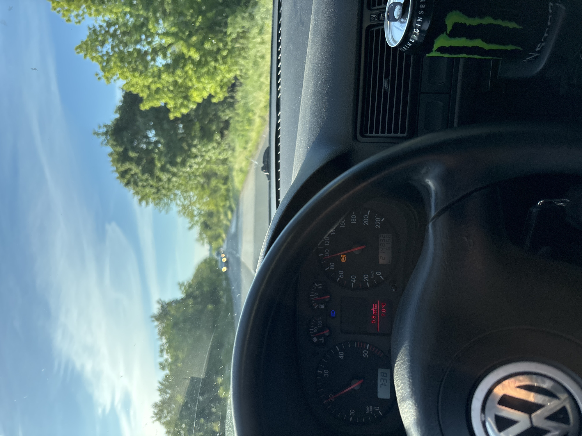 A view from the driver's seat of a car, focusing on the dashboard and the road ahead. The steering wheel, featuring the Volkswagen logo, dominates the foreground. The speedometer reads approximately 10 km/h, and the tachometer indicates 738 RPM. The temperature is shown as 7.0°C, and the fuel level appears to be at full. The odometer reads 214832. The dashboard includes an air vent with a partially visible can of Monster Energy drink. The view through the windshield reveals a road bordered by trees and a clear blue sky with some clouds. The location is Unknown, Unknown.