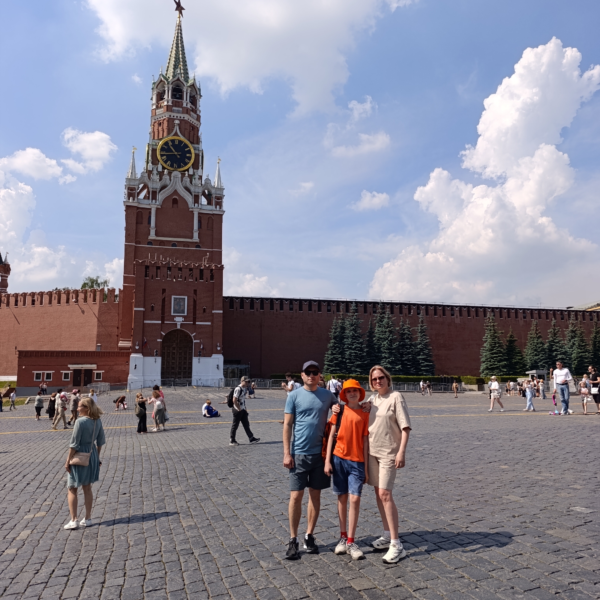 The image depicts a family of three posing for a photo in front of a prominent clock tower and a red brick wall, likely a historical landmark. The family consists of a man wearing a blue t-shirt and dark shorts, a boy in an orange t-shirt and blue shorts with an orange hat, and a woman in a beige t-shirt and shorts. They are standing on a cobblestone ground in an open square. In the background, other people are visible walking around and observing the surroundings. The clock tower features a large clock face with Roman numerals and a red star at its apex. Tall, dark green evergreen trees are planted in a line along the base of the red brick wall. The sky is blue with scattered white clouds. The location is identified as Алабино, Russia.