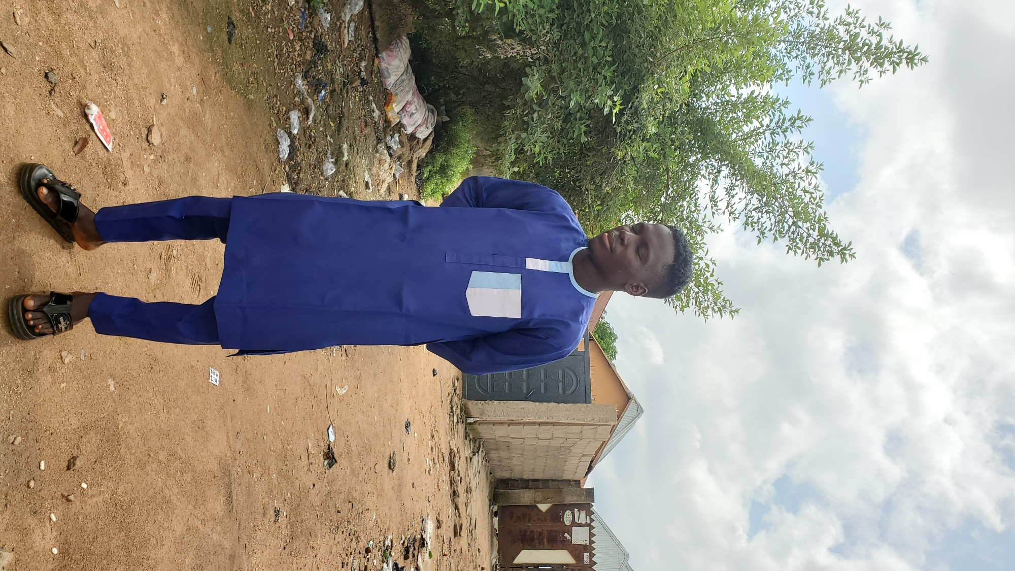 A young Black male stands in a dirt alleyway in Jalingo, Nigeria. He is wearing a blue traditional outfit with a light blue and white patterned pocket and trim. His hands are clasped behind his back, and his eyes are closed. He is wearing black flip-flops. The background shows a leafy green tree on the left and a row of buildings with corrugated metal roofs and brick walls on the right. The ground is covered in dirt, debris, and scattered trash, including plastic bags and wrappers. The sky is overcast with scattered clouds.